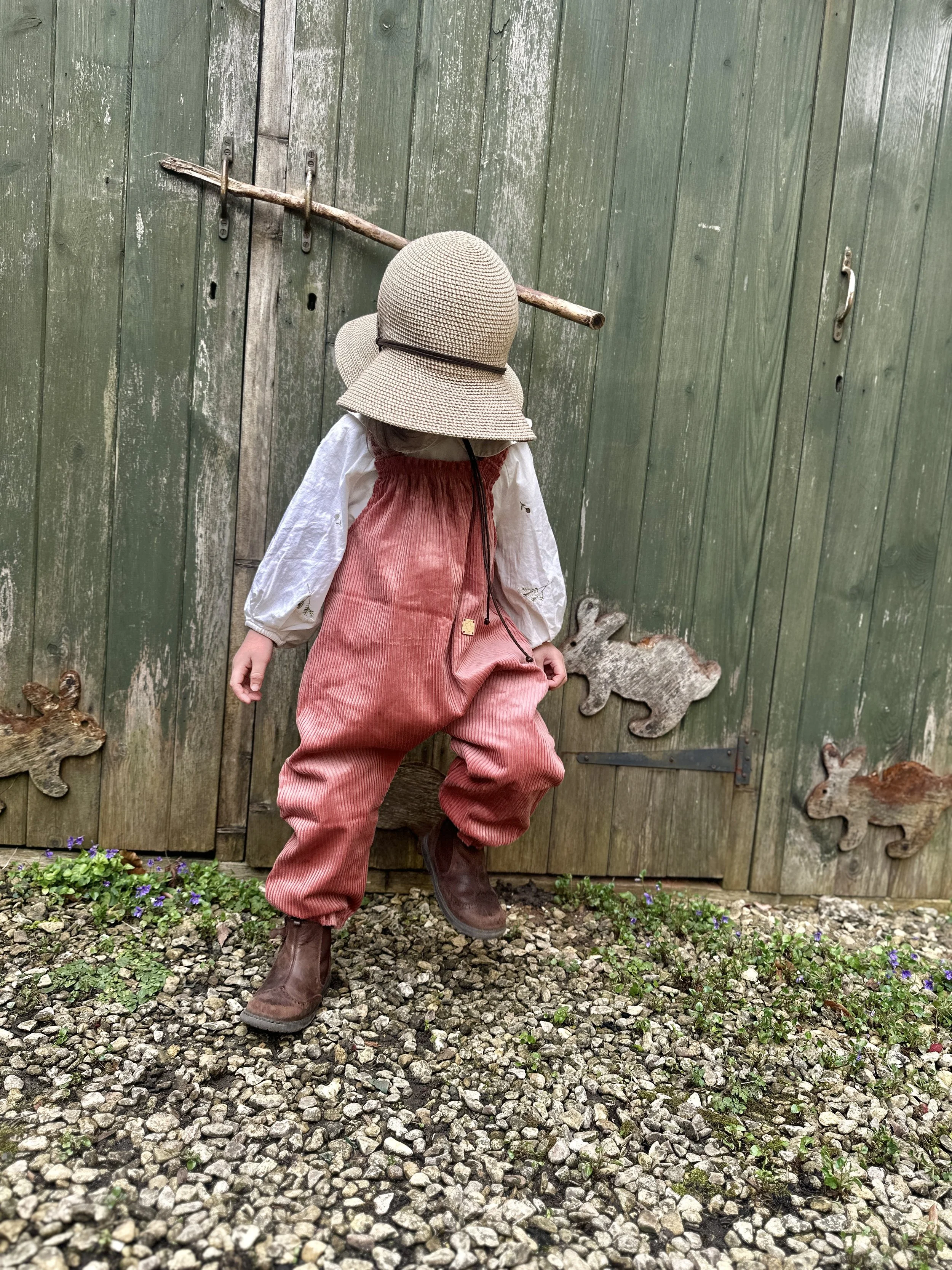child wearing organic cotton corduroy pullover dungarees in coral colour, brown boost, sunhat, standing in front a wooden shed