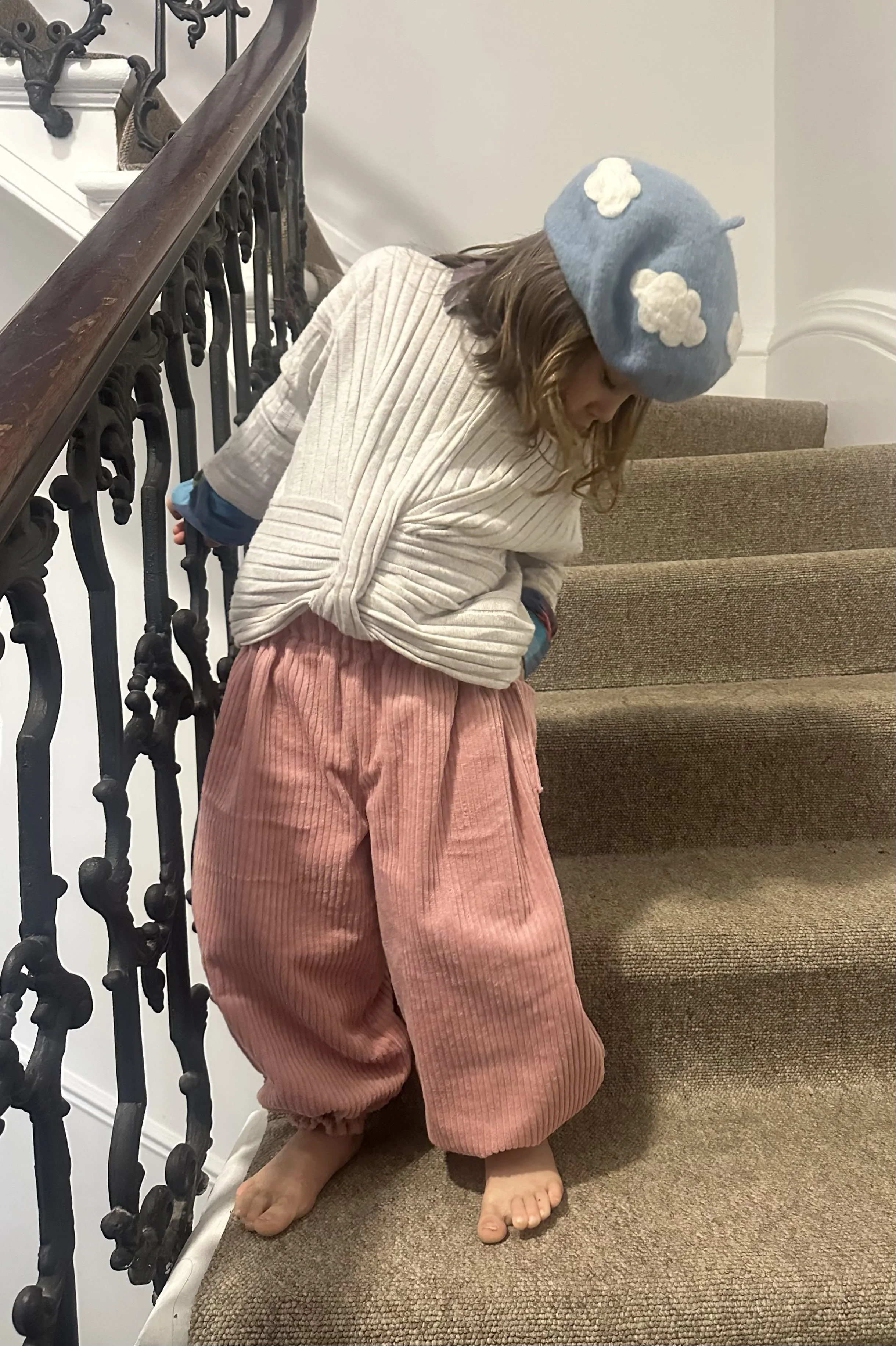 child wearing organic cotton corduroy pink trousers barrel shape baggy fit oversized elastic hem, with white top, blue cloudy hat, standing on staircase