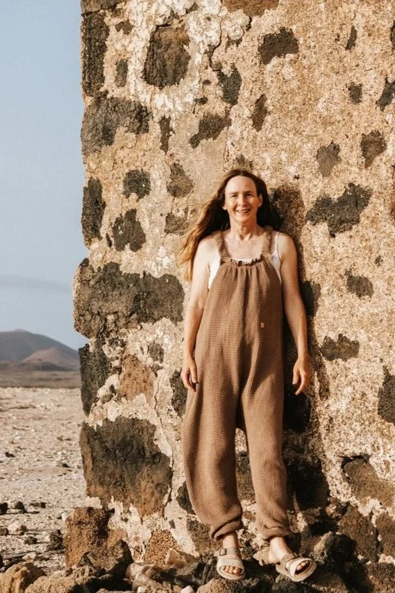 woman wearing organic cotton waffle pullover dungarees beige colour over a white t shirt, standing againt a cliff wall on the sea side