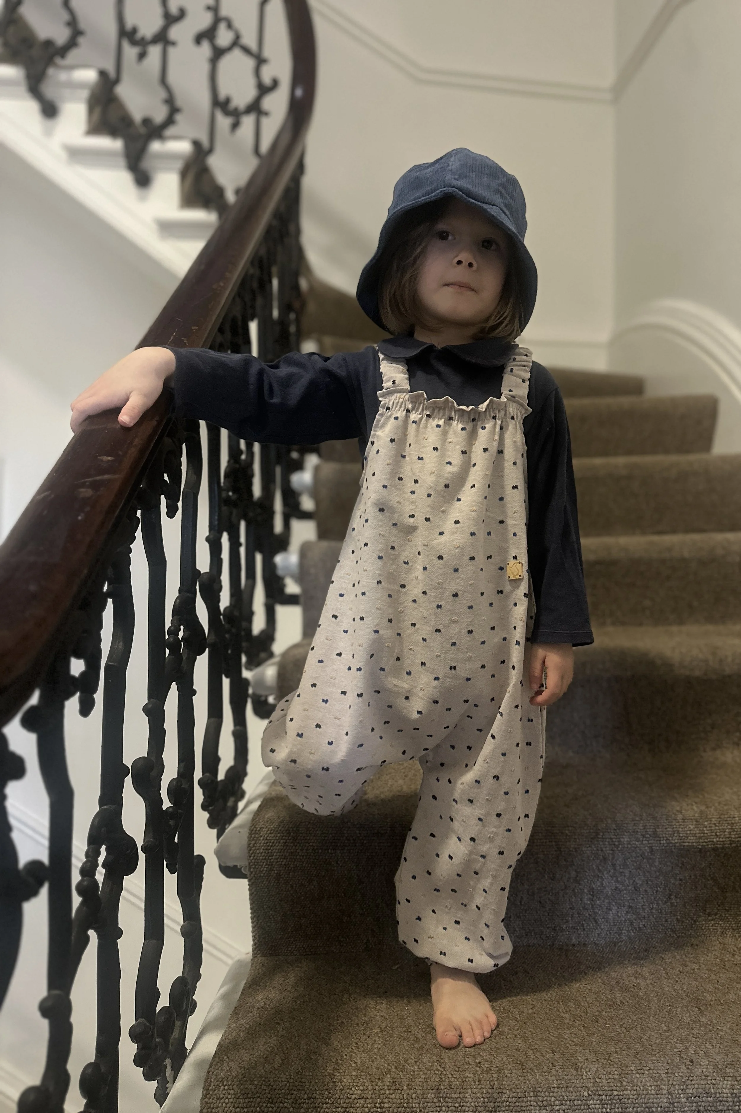 child wearing organic cotton speckled dungarees Neutral colour, layered over a navy top, and a navy bucket hat