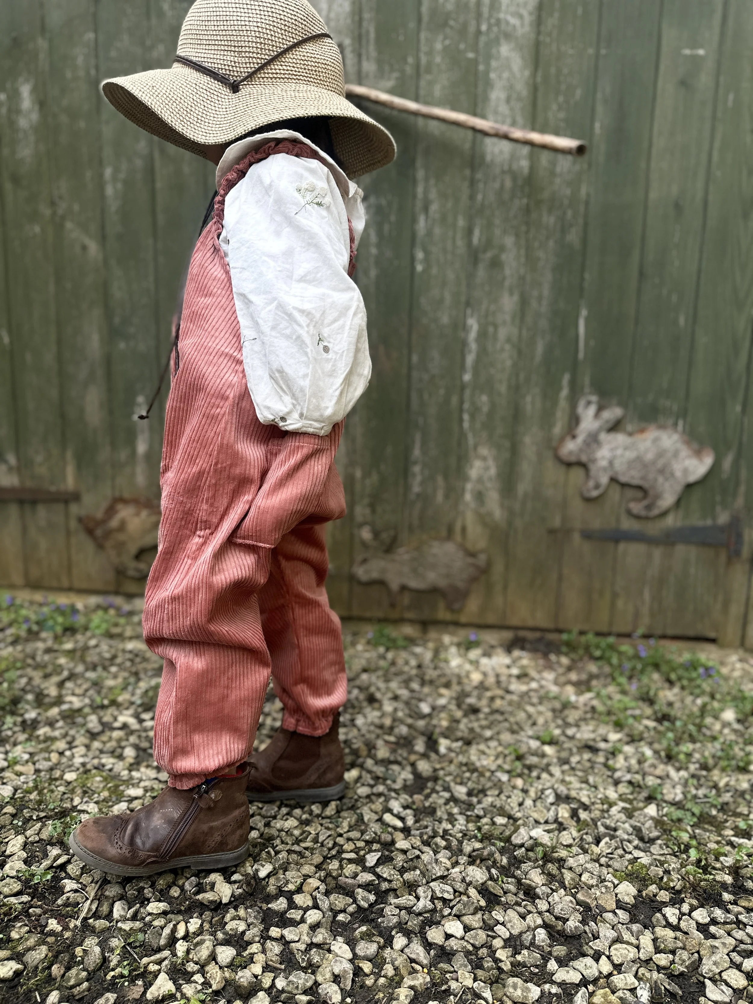 side view of child wearing organic cotton corduroy pullover dungarees in coral colour, brown boost, sunhat, standing in front a wooden shed