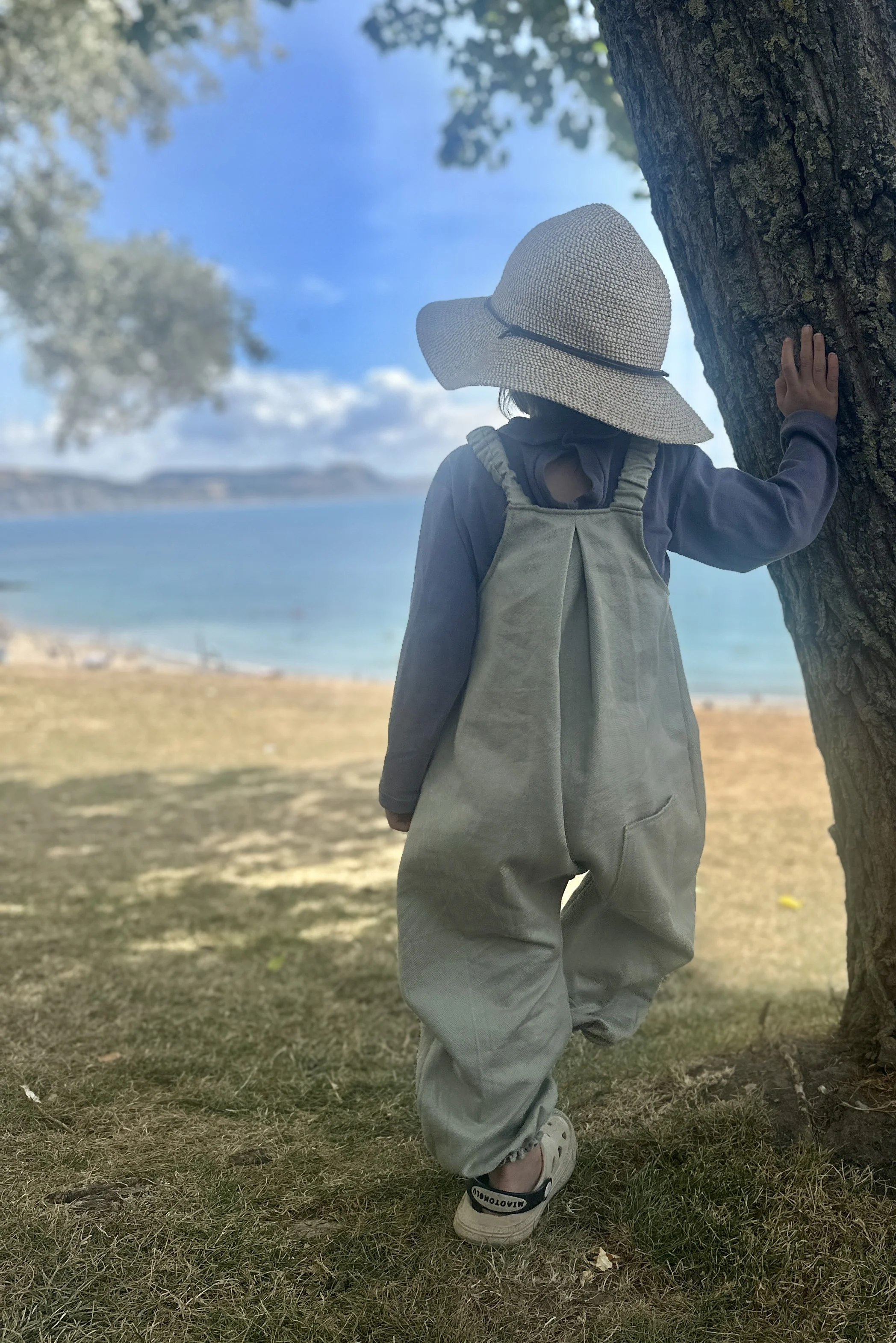 rear view of child wearing organic cotton denim light sage green pullover dungarees layered with purple top, sun hat, standing under a tree on the coast