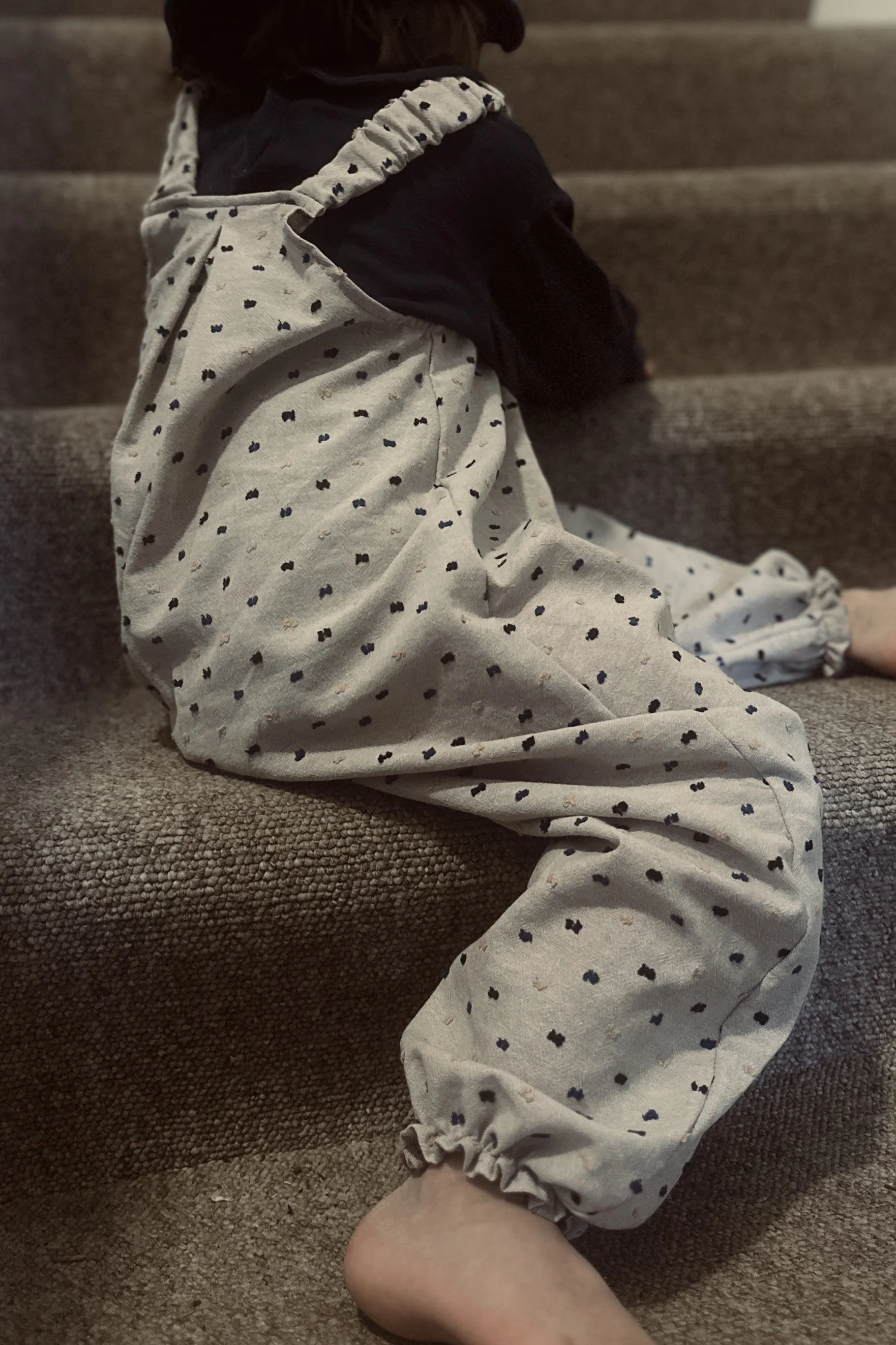 rear of child wearing organic cotton speckled dungarees Neutral colour, layered over a navy top, and a navy bucket hat