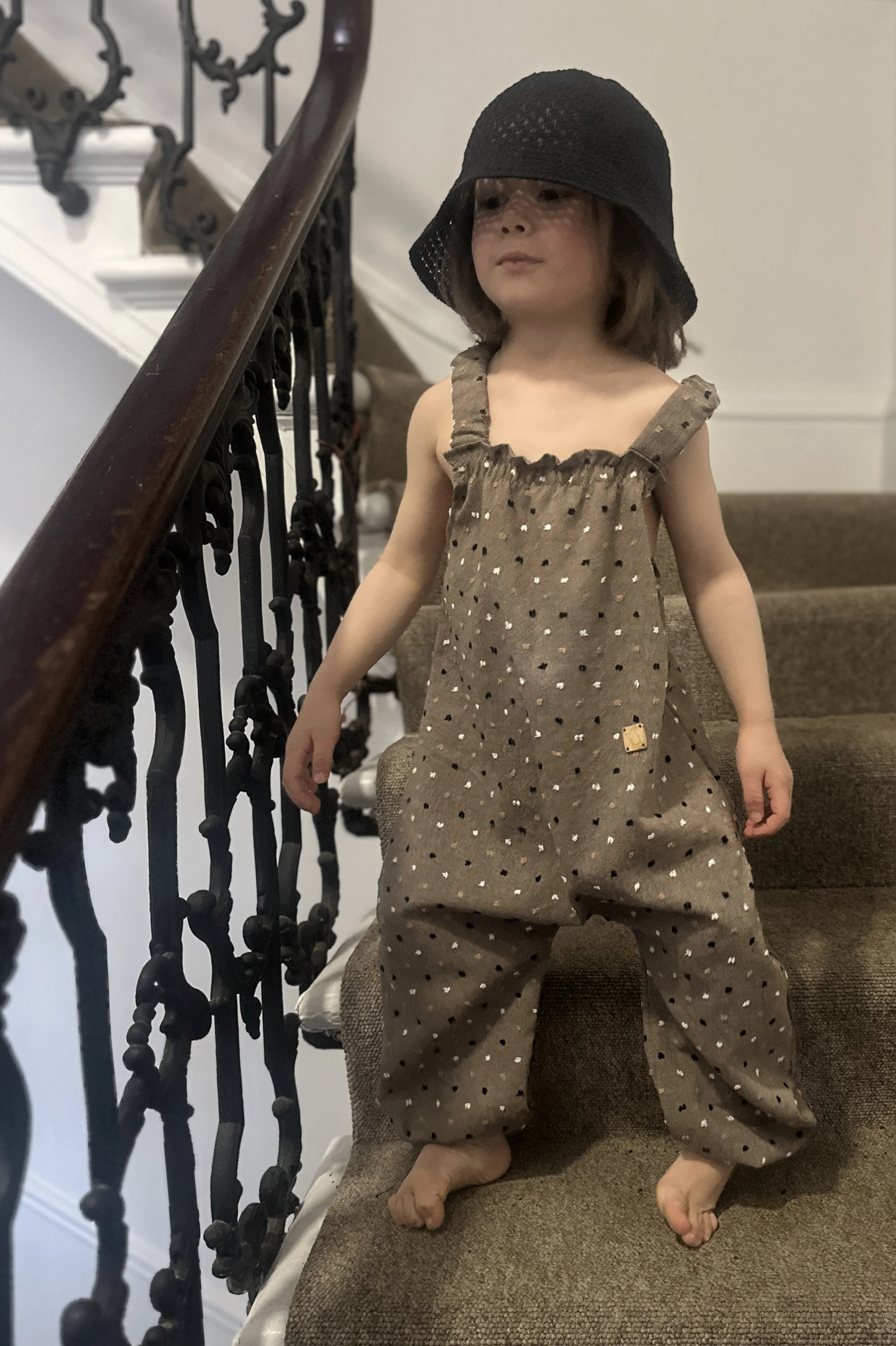 child wearing organic cotton speckled dungarees in cocoa colour, with a black sun hat, standing on staircase