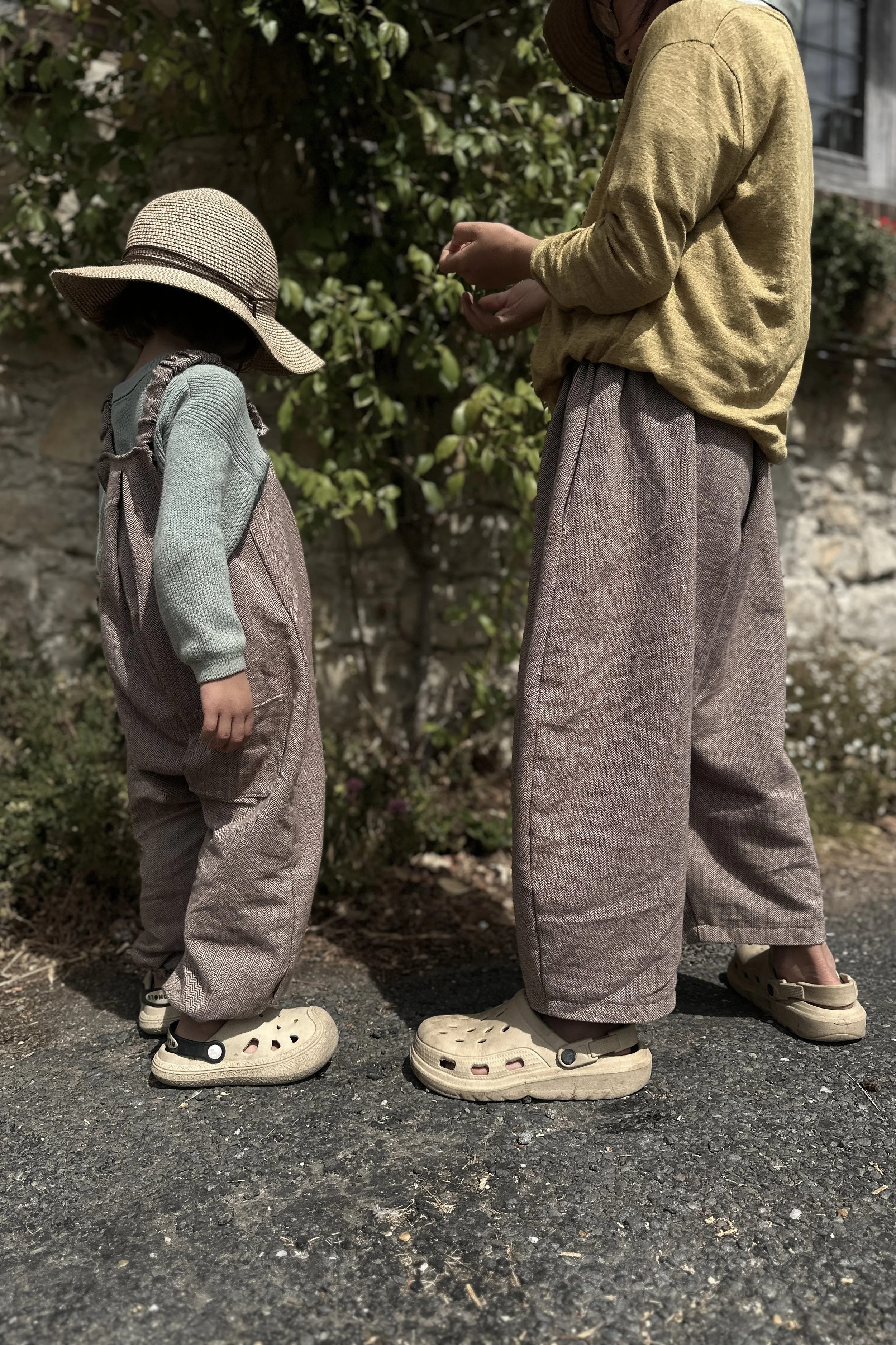 adult wearing organic cotton herringbone brown barrel leg trousers full length, standing with a child wearing wearing organic cotton herringbone brown dungarees