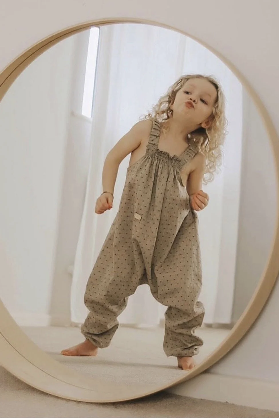 child wearing organic cotton pullover dungarees dotty beige, standing in front of big round mirror