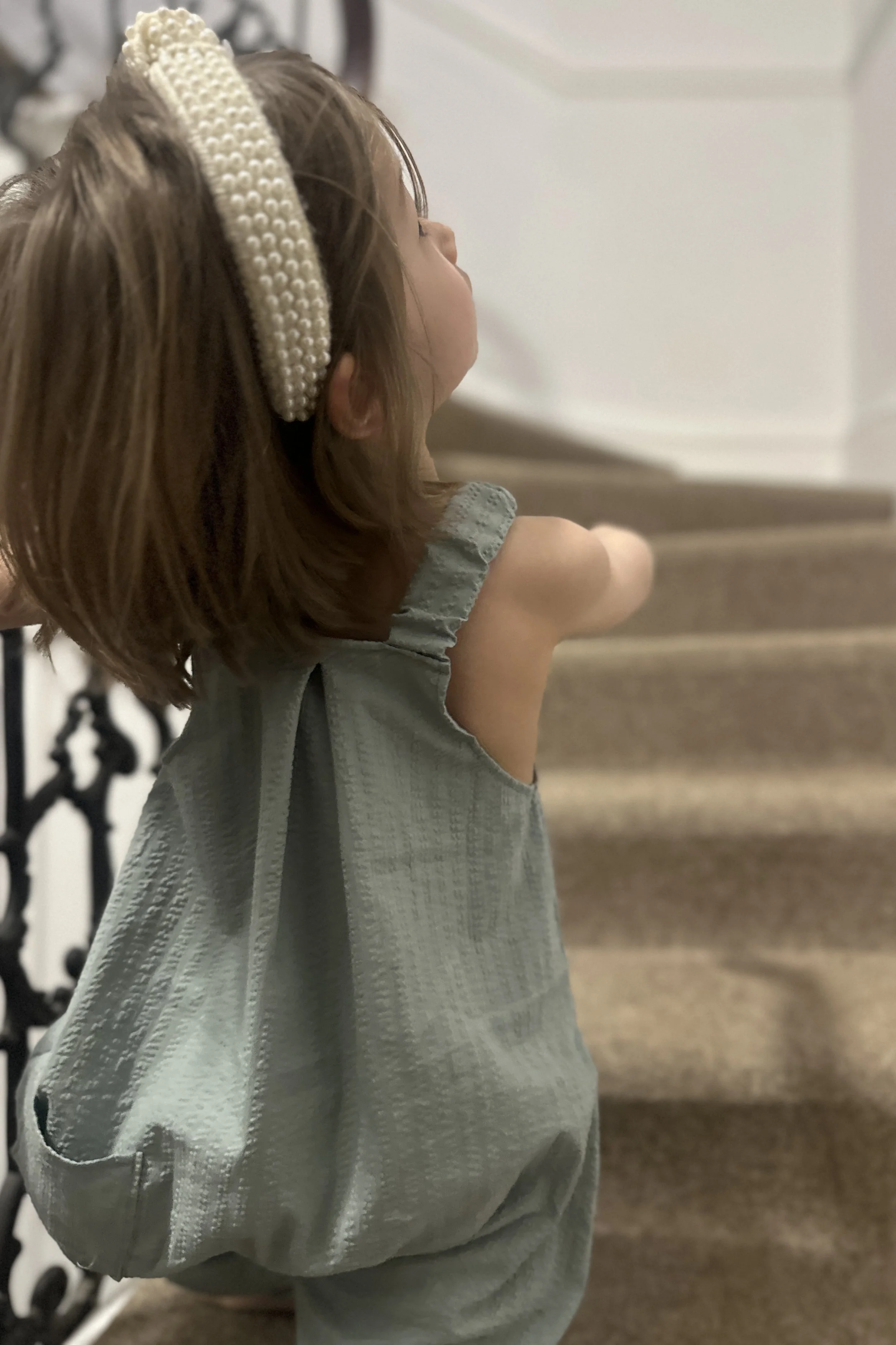 rear of Child wearing organic cotton seersucker Duck Egg Blue pullover dungarees, pearl headband, sitting on stairs