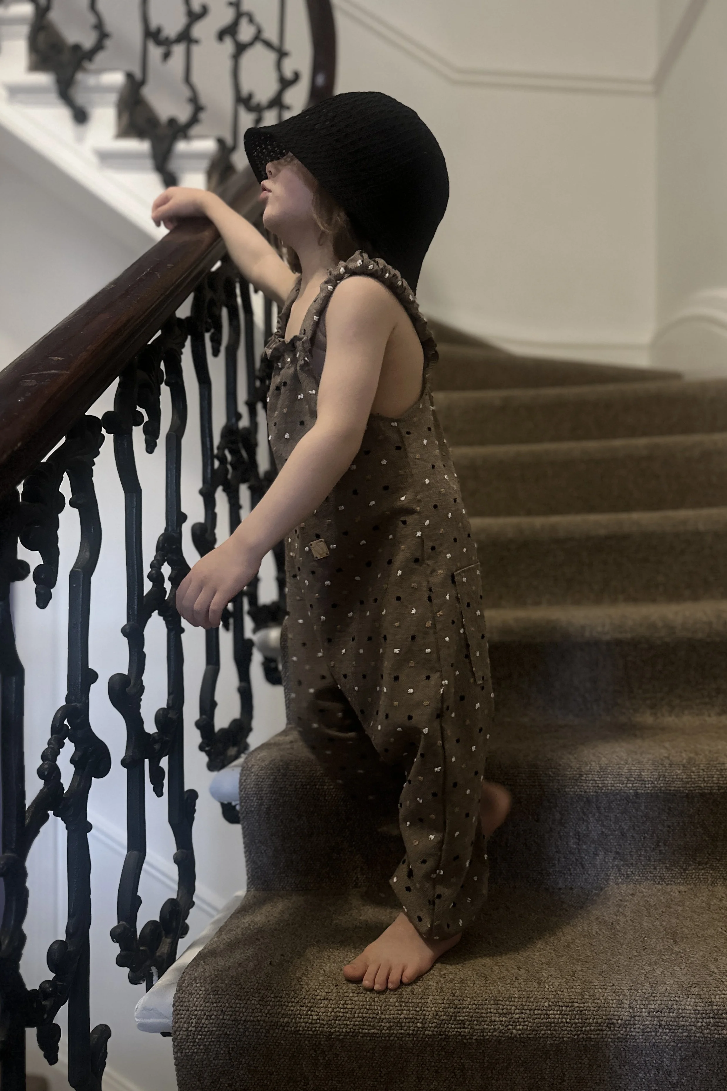 side view of child wearing organic cotton speckled dungarees in cocoa colour, with a black sun hat, standing on staircase