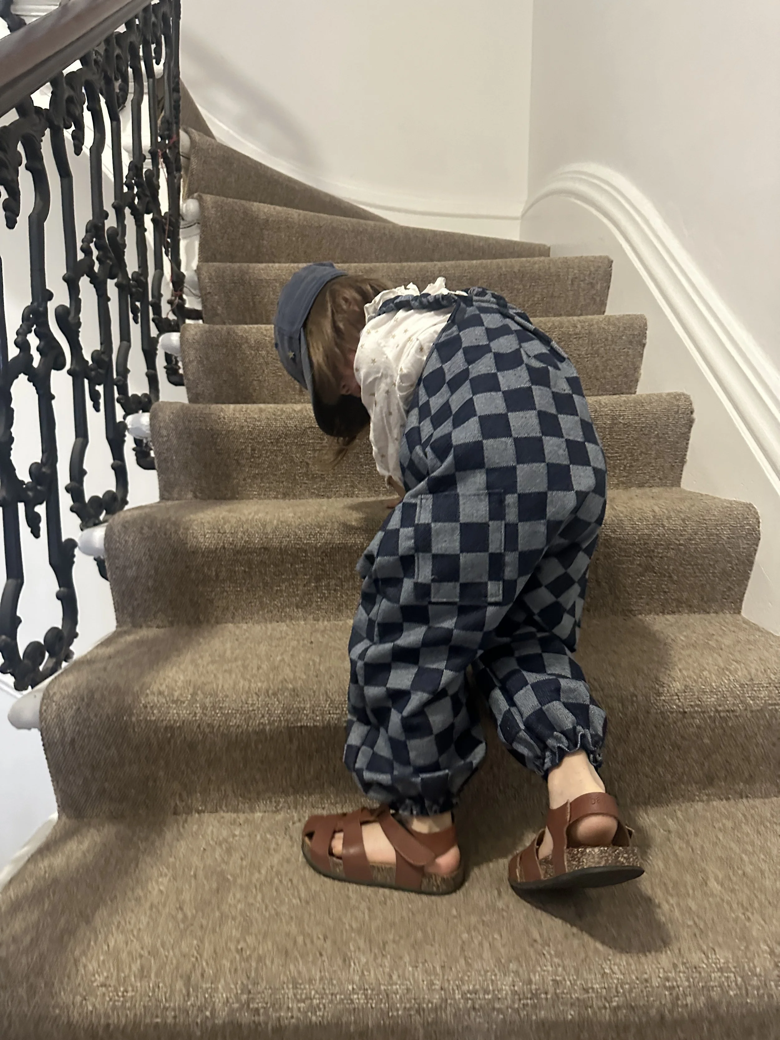 back view of child wearing organic cotton checkerboard denim blue dungarees, brown sandals, denim cap, standing on staircase, highlight pocket at the back