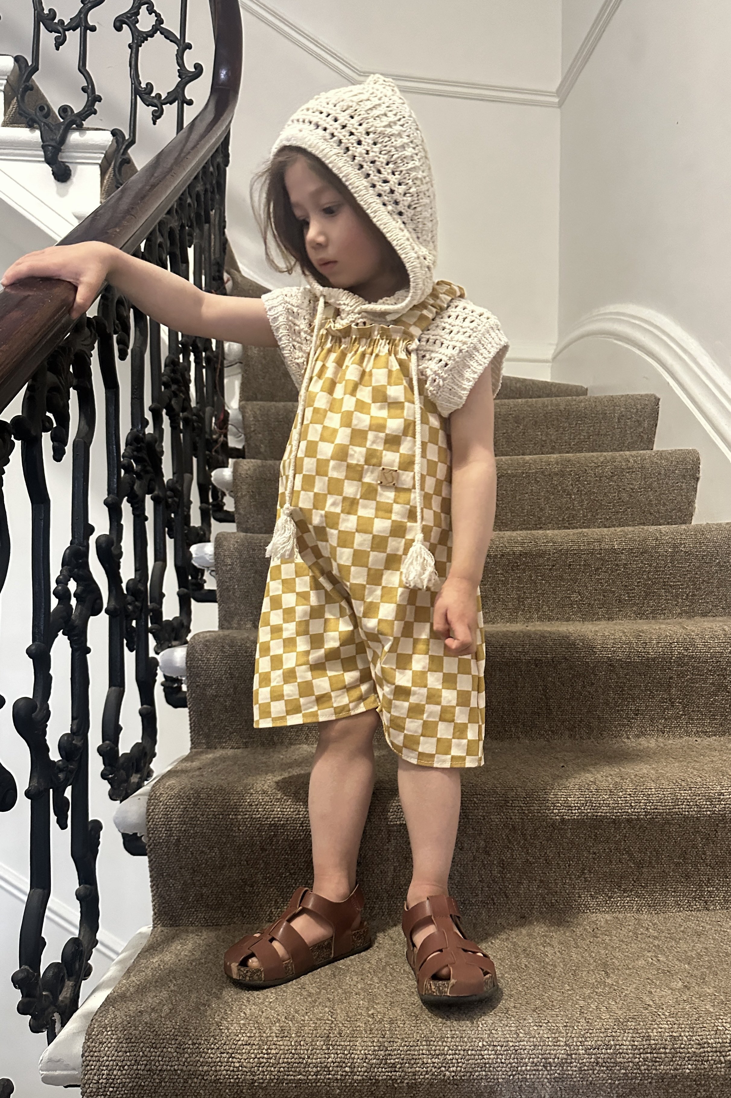 child wearing organic cotton canvas pullover short dungarees in mustard checkerboard colour, layered over a crochet short sleeves hoodie top, brown sandals