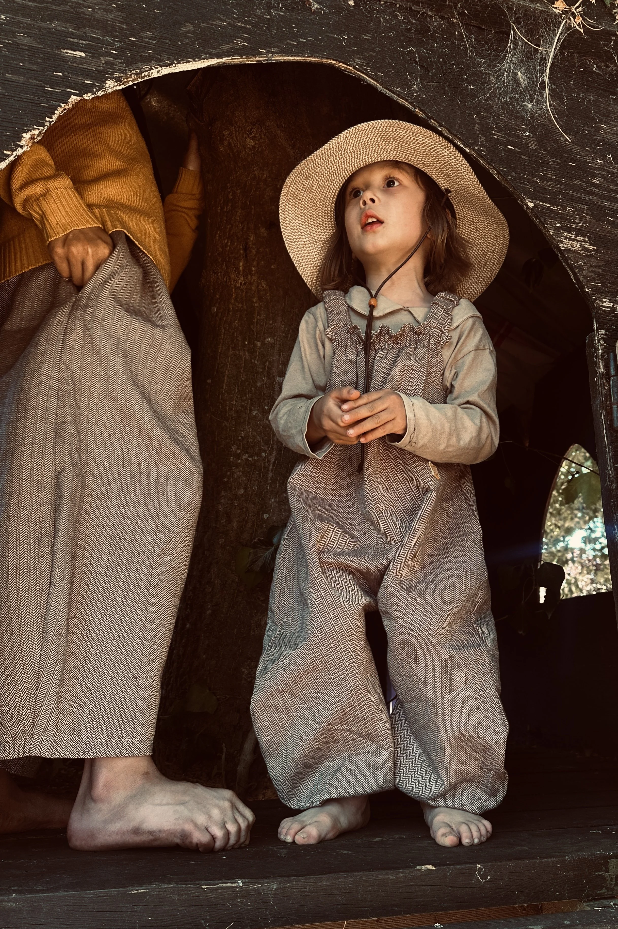 child wearing organic cotton herringbone brown dungarees, layered with sage green jumper, sunhat, standing in a tree house next to big brother wearing barrel leg trousers made of the same fabric