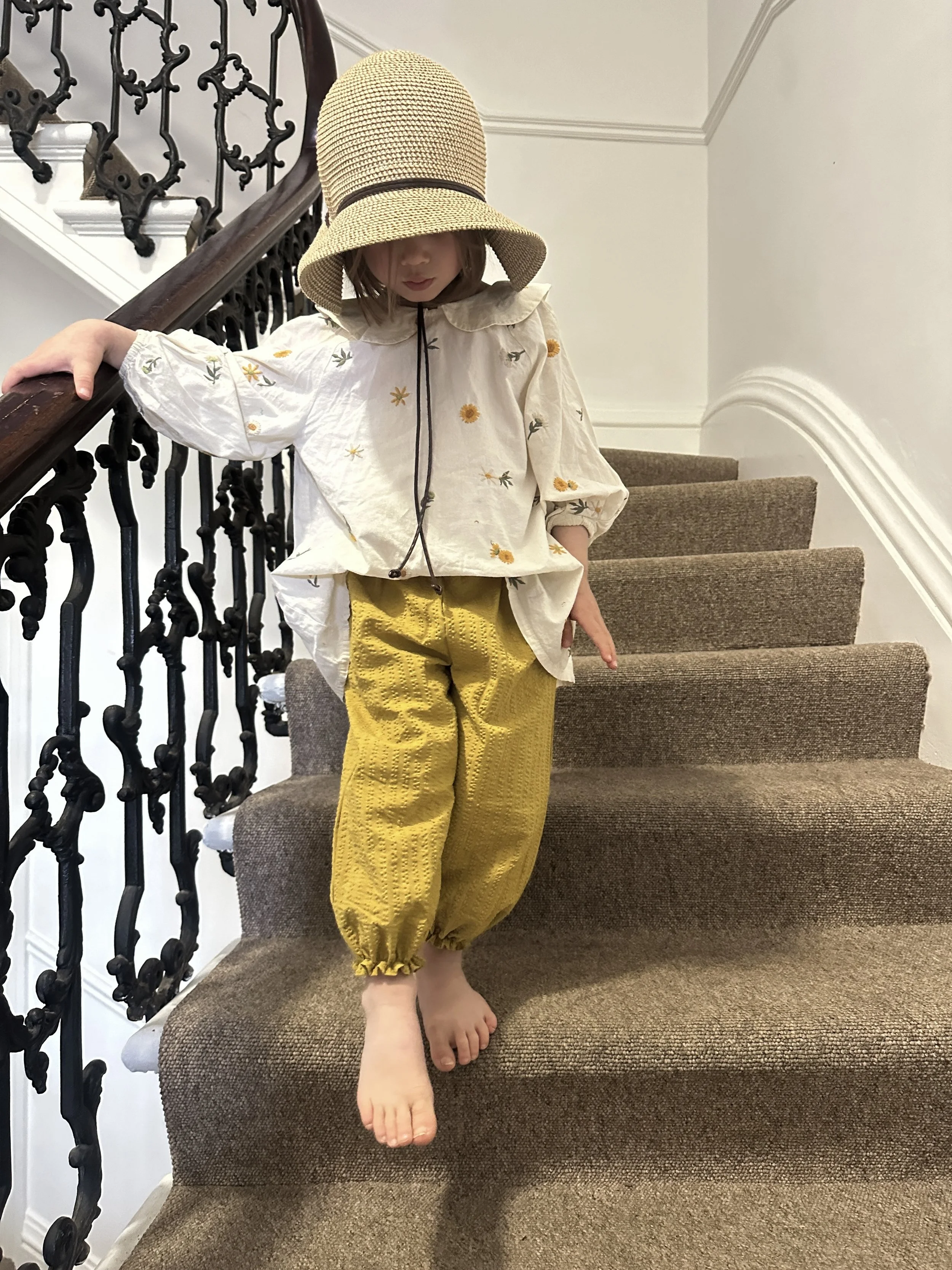 child wearing organic cotton seersucker barrel trousers mustard colour, paired with white blouse with Peter Pan collar and embroidered flowers, sun hat
