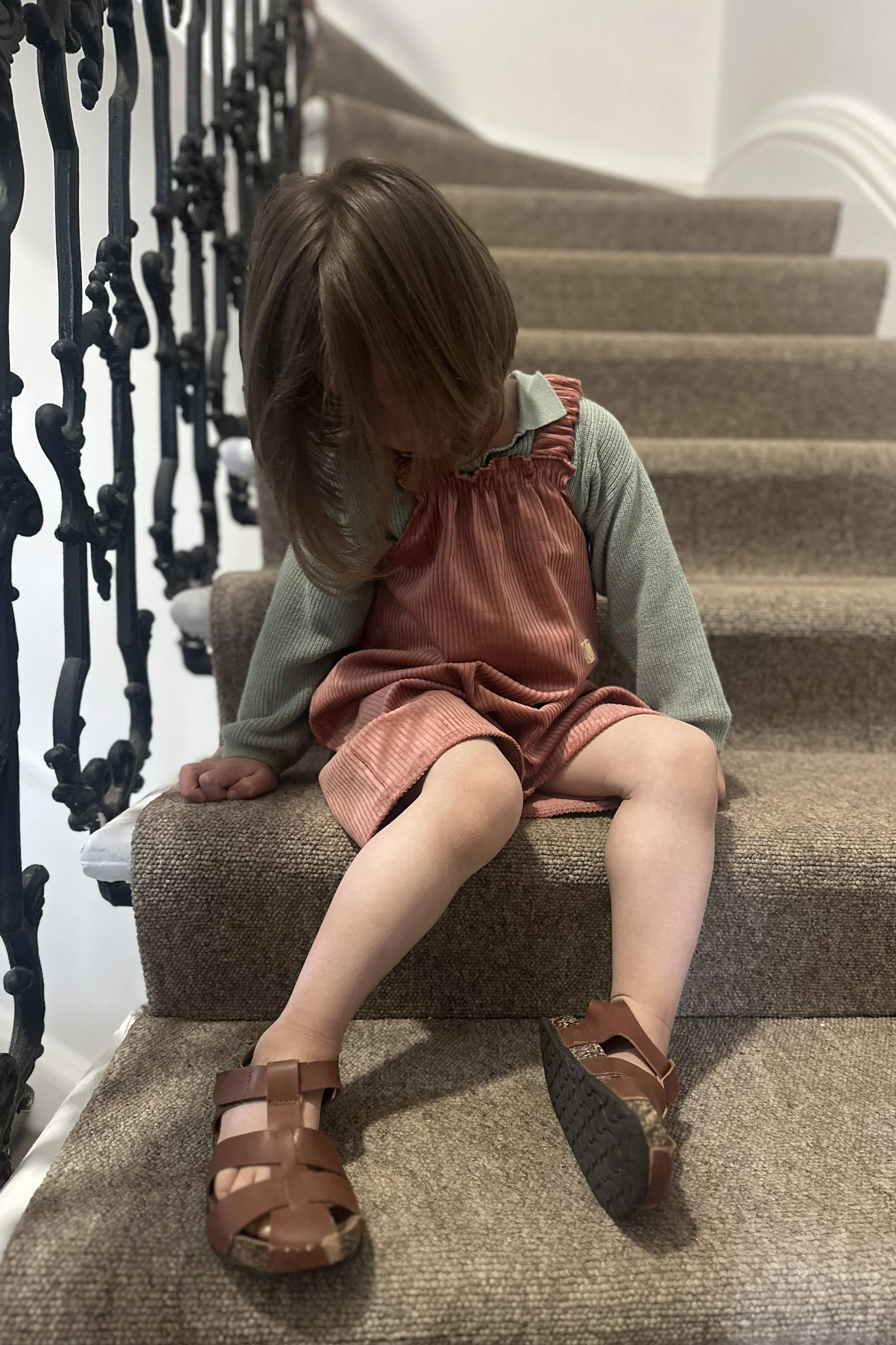 child wearing organic cotton corduroy short dungarees coral colour, layered over a duck egg green jumper, brown sandals, sitting on staircase
