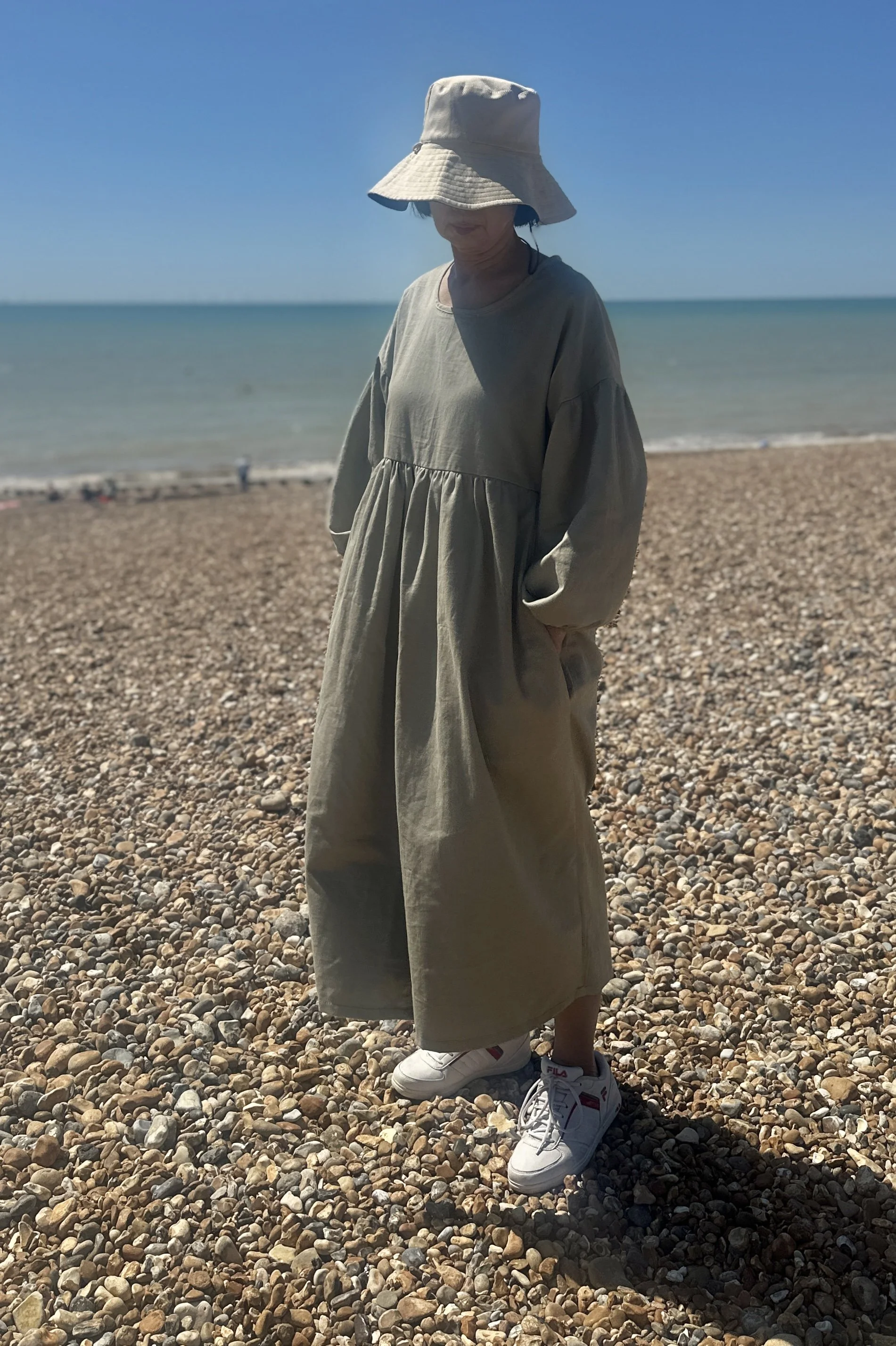adult wearing organic cotton denim light sage maxi dress, hand in pocket, white trainers, beige sunhat, walking on the beach