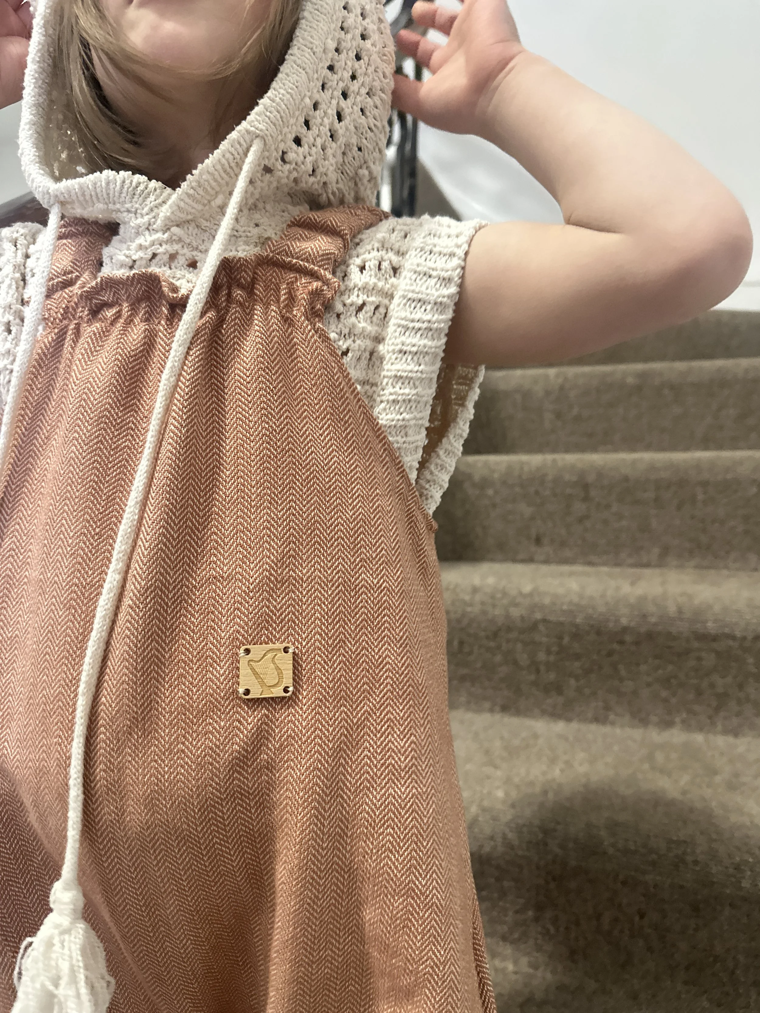 close up child wearing organic cotton herringbone short dungarees peach colour, layered over a cream knitted short sleeves hoodie, paired with brown sandals, standing on staircase