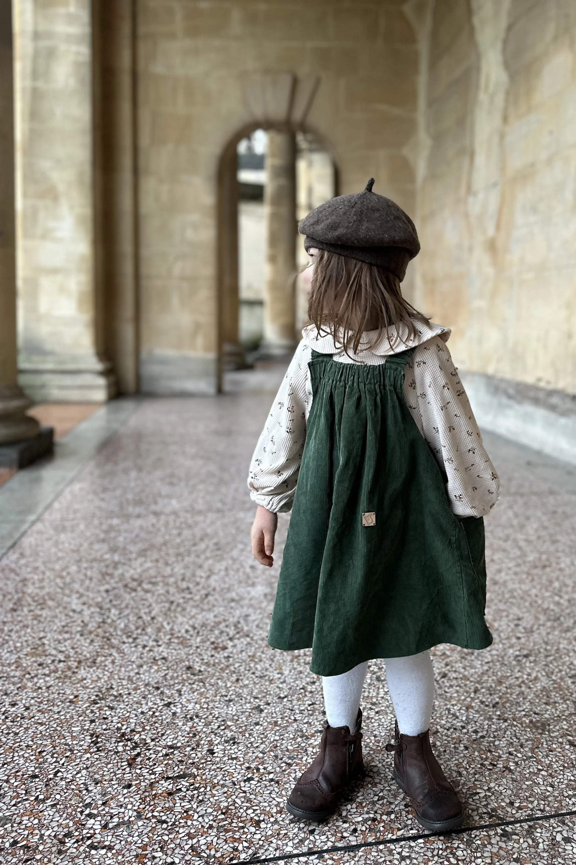 child wearing organic cotton needlecord dress dark green, layered over floral blouse with Peter Pan collar, a brown woolly hat and brown boots, highlighting pocket at the back