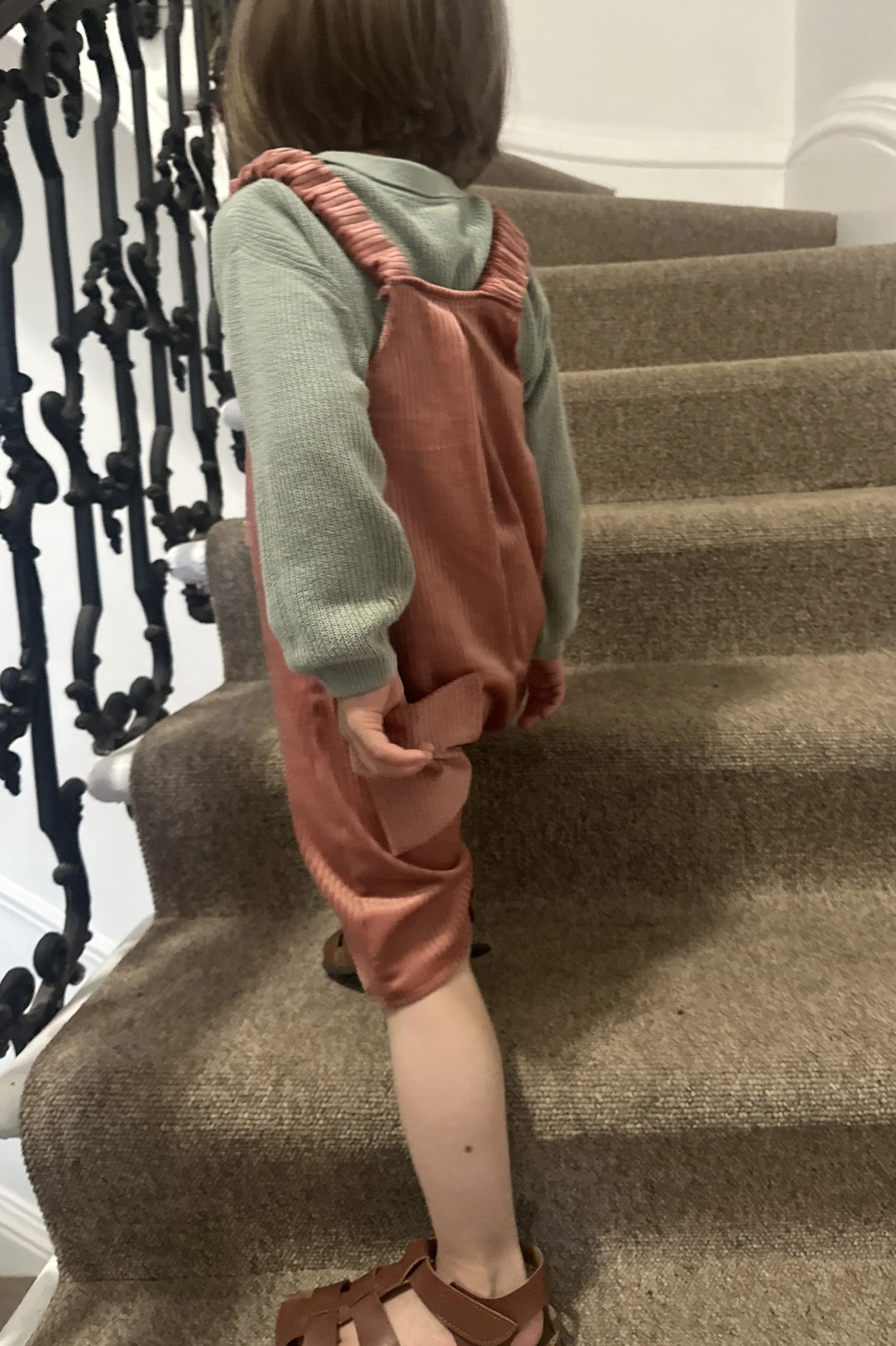 back view of child wearing organic cotton corduroy short dungarees coral colour, layered over a duck egg green jumper, brown sandals, standing on staircase