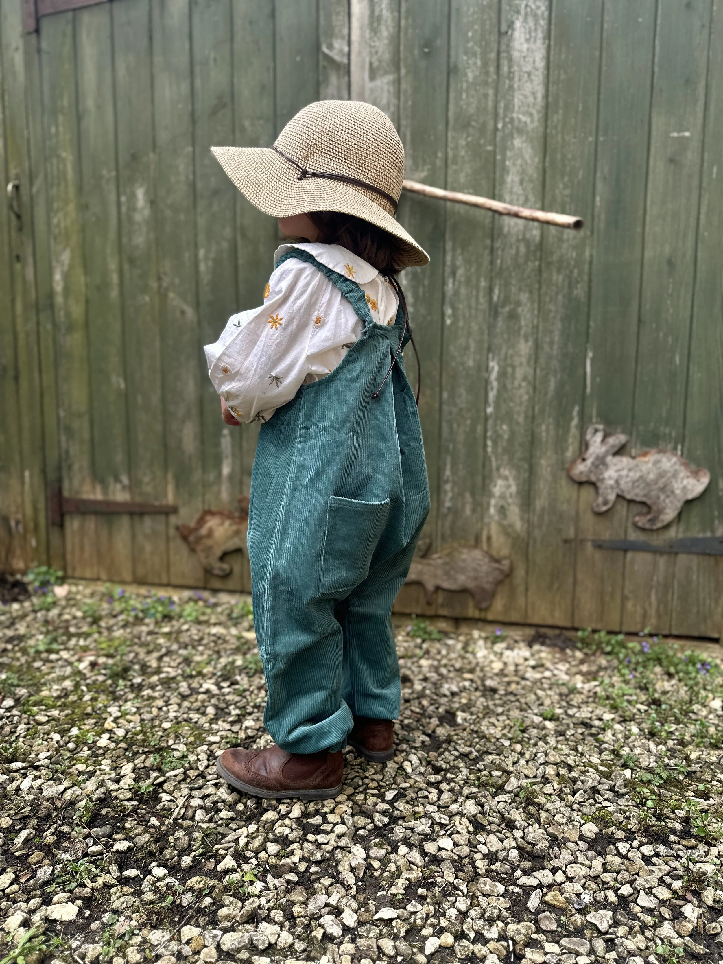 back view of child wearing organic cotton corduroy teal green pullover dungarees, brown boots, sun hat, standing in front of wooden shed