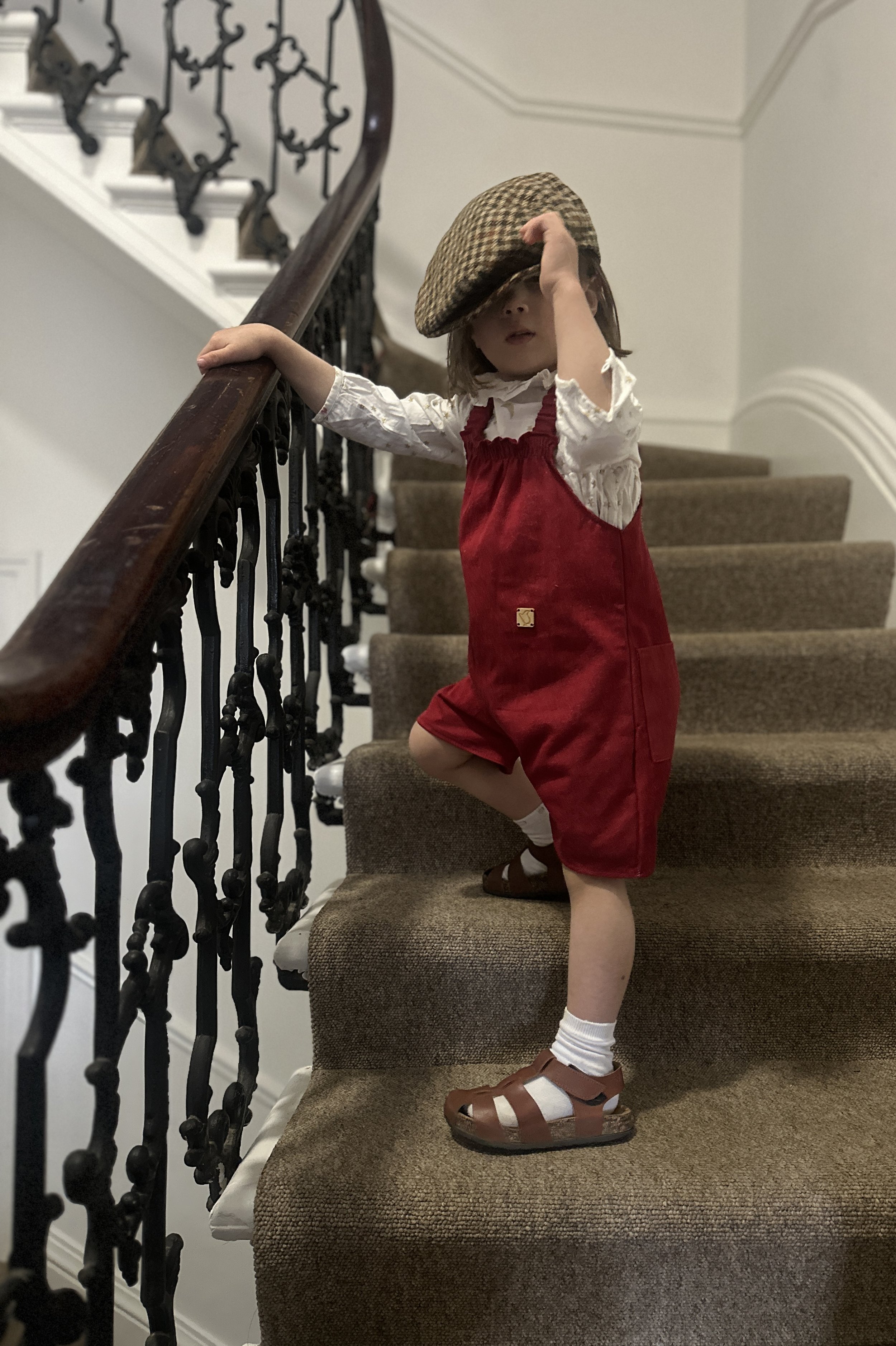 child wearing organic cotton denim red short dungarees over white blouse with golden stars with brown sandals tartan hat, standing on staircase