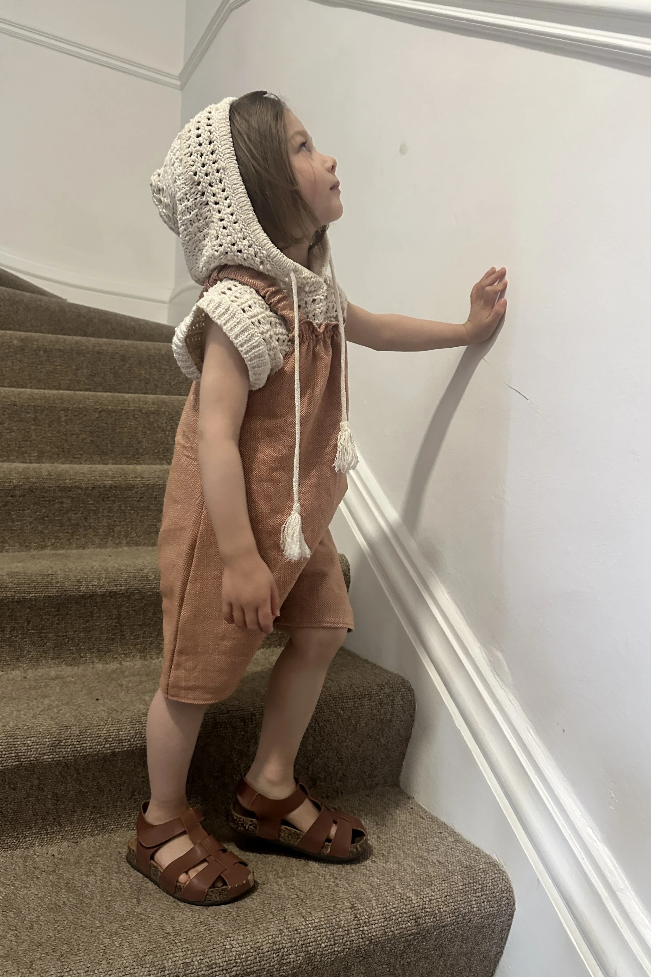 child wearing organic cotton herringbone short dungarees peach colour, layered over a cream knitted short sleeves hoodie, paired with brown sandals, standing on staircase