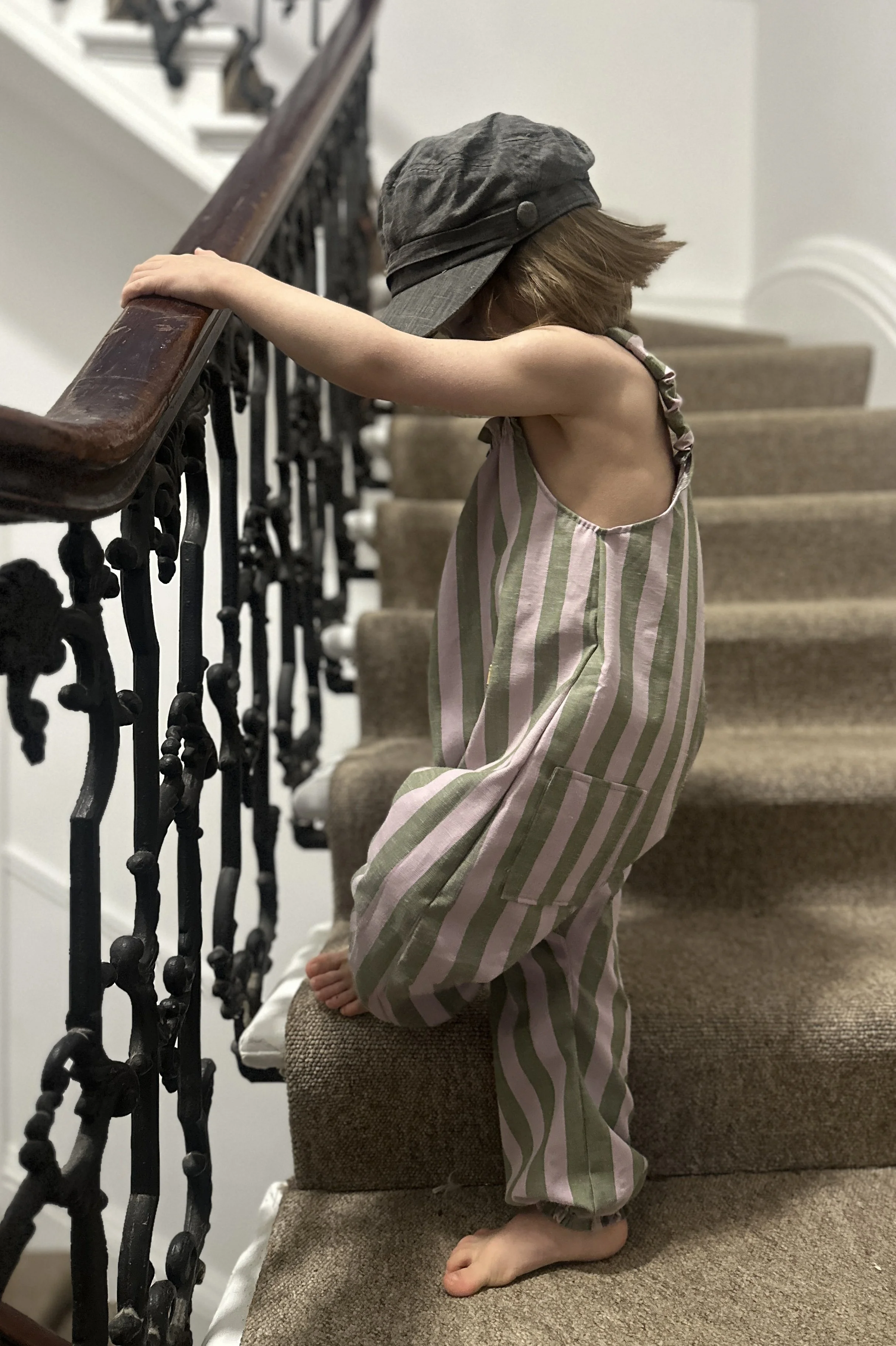 side view of Child wearing organic cotton and linen dungarees in wide stripe olive green and lilac, and a grey hat, standing on staircase (Copy)