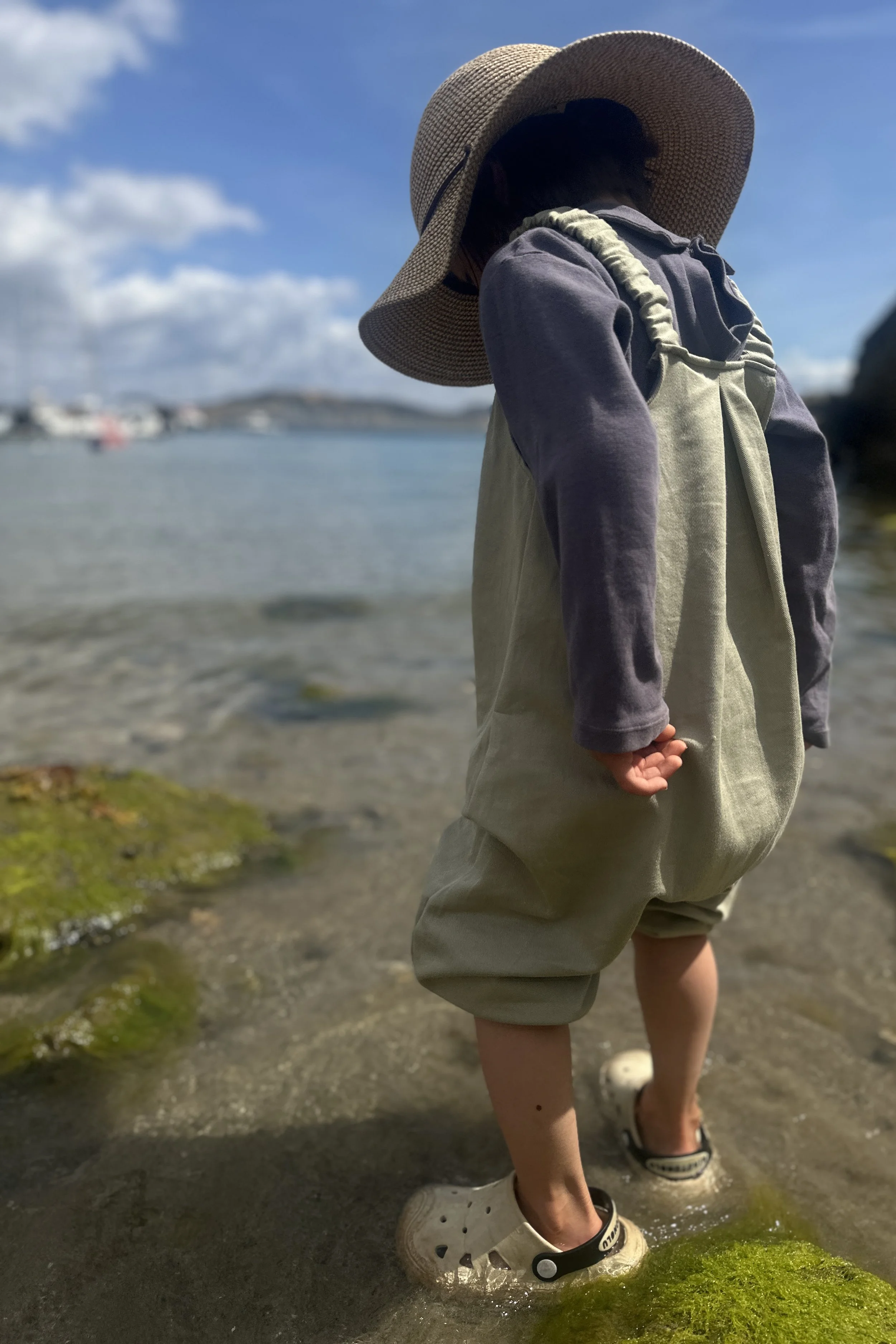 rear view of child wearing organic cotton denim light sage green pullover dungarees layered with purple top, sun hat, standing in the sea