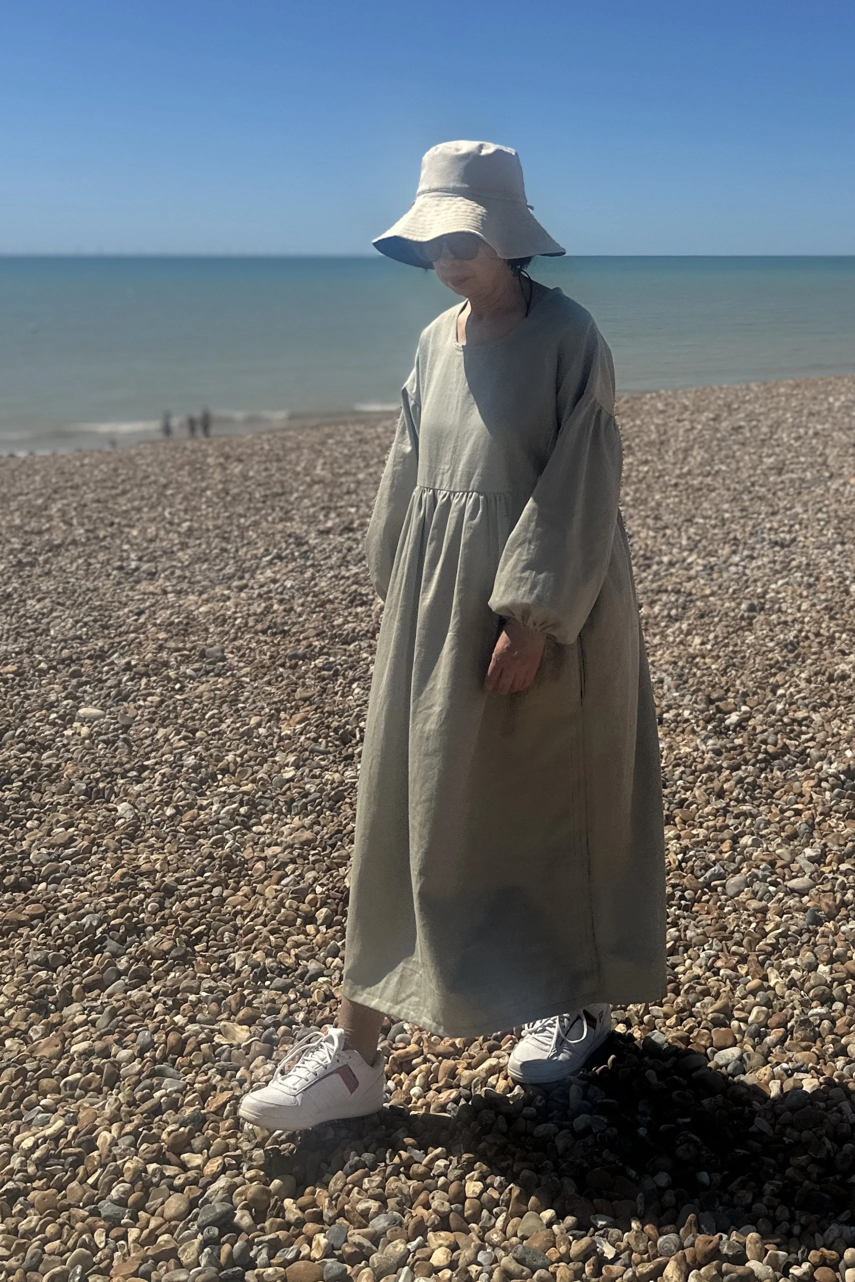 adult wearing organic cotton denim light sage maxi dress, white trainers, beige sunhat, walking on the beach