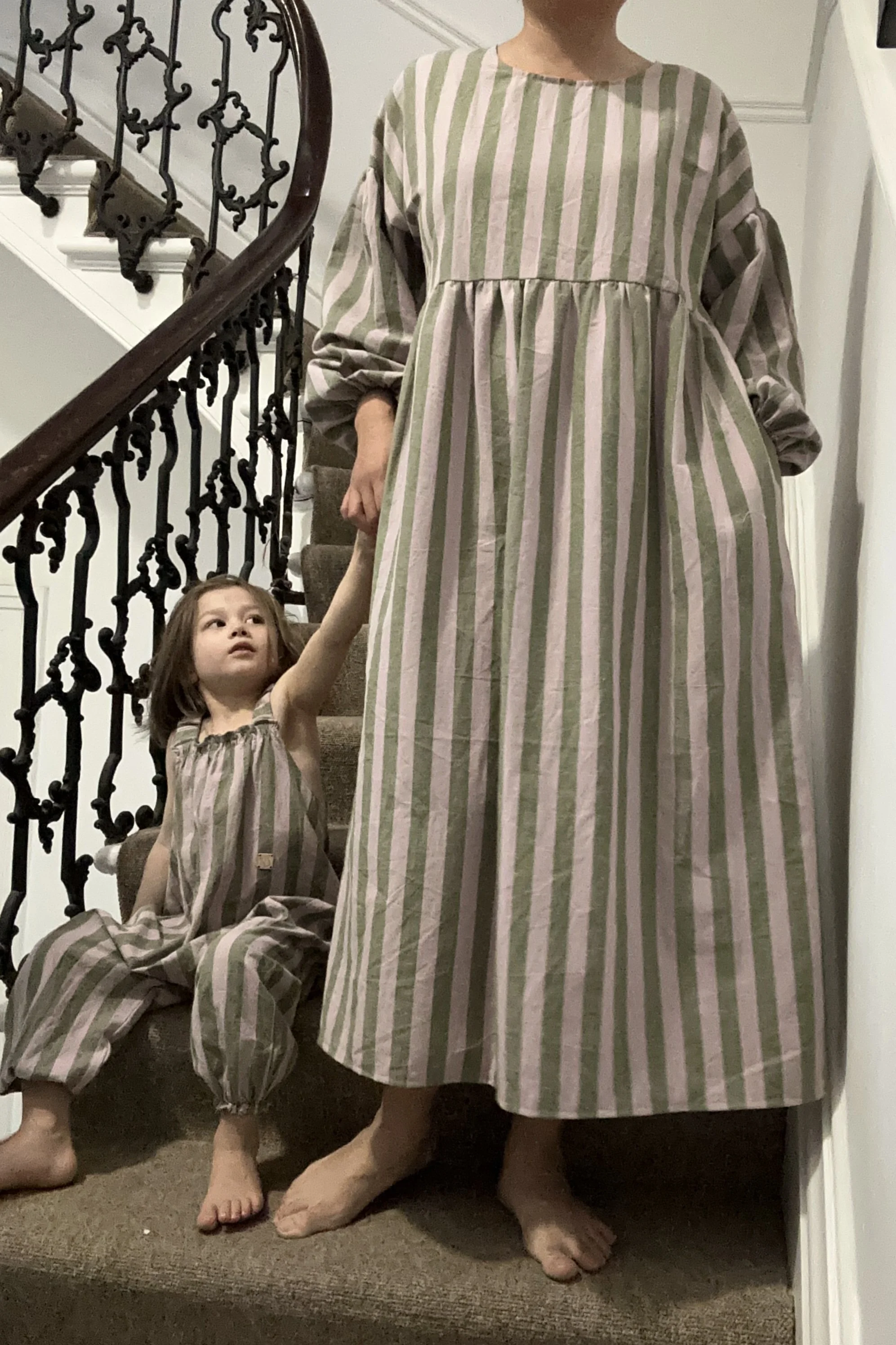 adult wearing organic cotton linen stripe olive green & lilac full length dress with balloon sleeves, with child wearing dungarees made of same fabric sitting next to her on staircase
