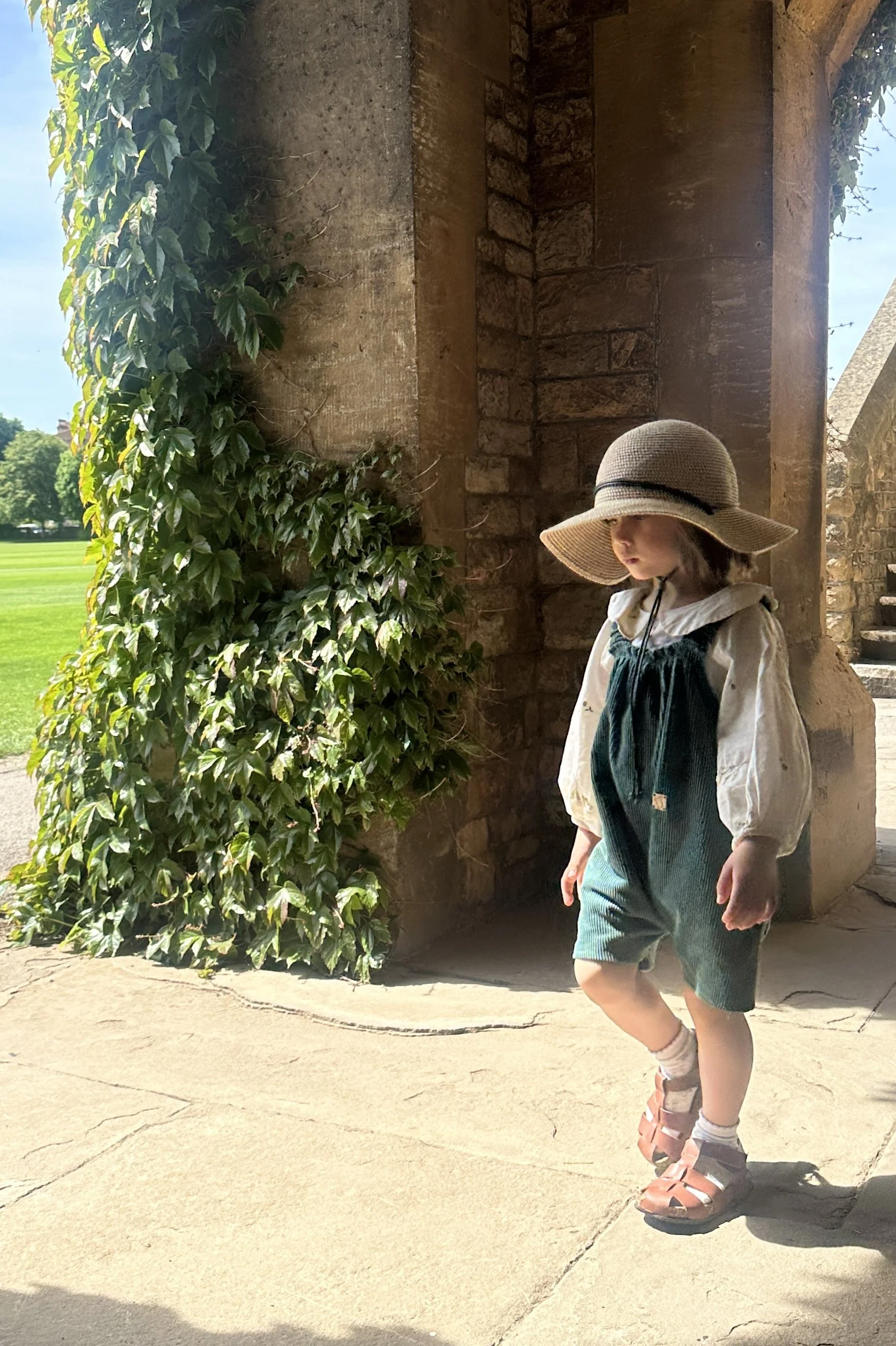 Child wearing teal green organic cotton corduroy short dungarees styled with a white embroidered blouse, sun hat, and brown sandals, standing outdoors under a stone archway in warm sunlight with a green natural backdrop