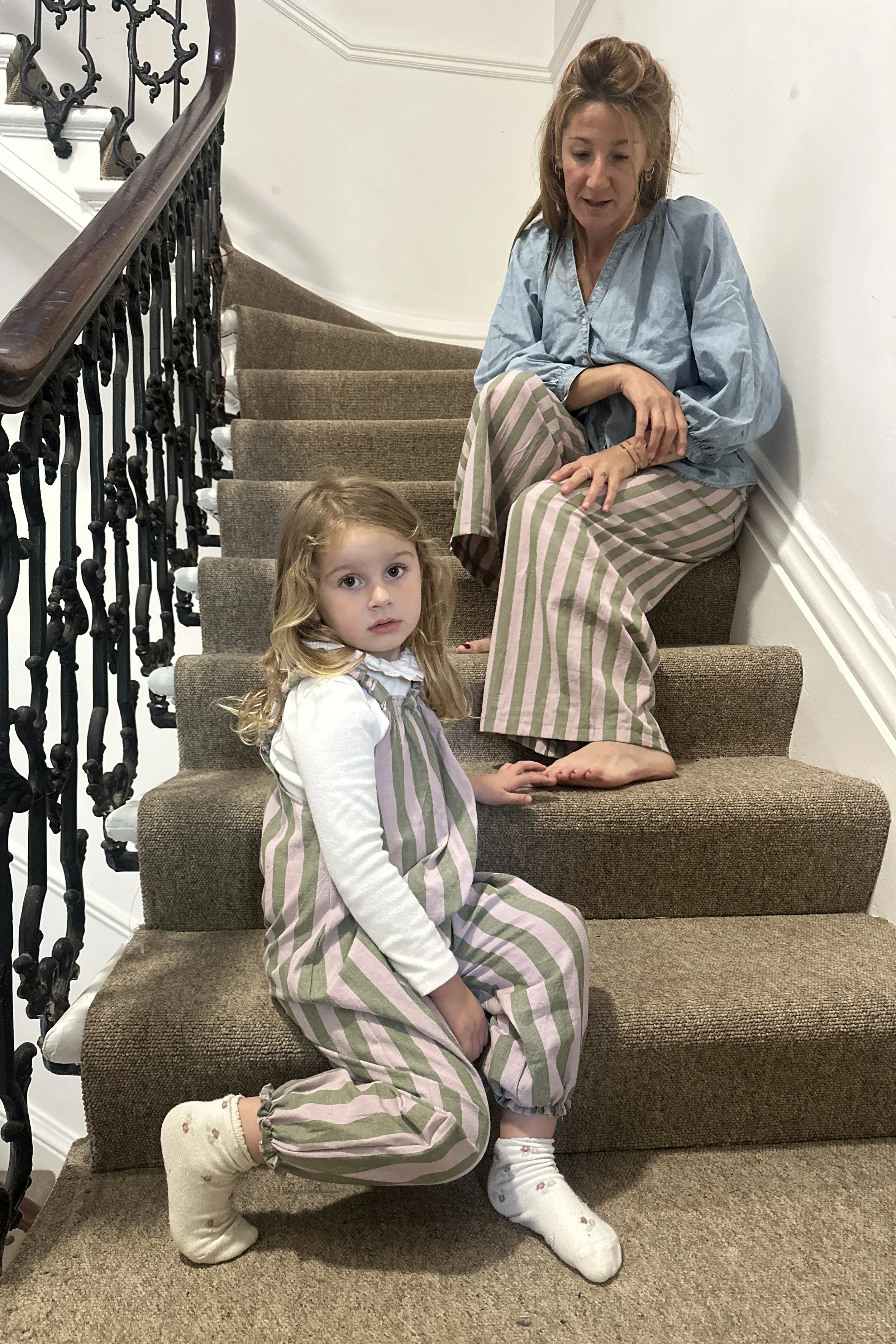 child wearing organic cotton linen sage and lilac stripe dungarees, mum wearing wide leg trousers with same fabric sitting on the stairs
