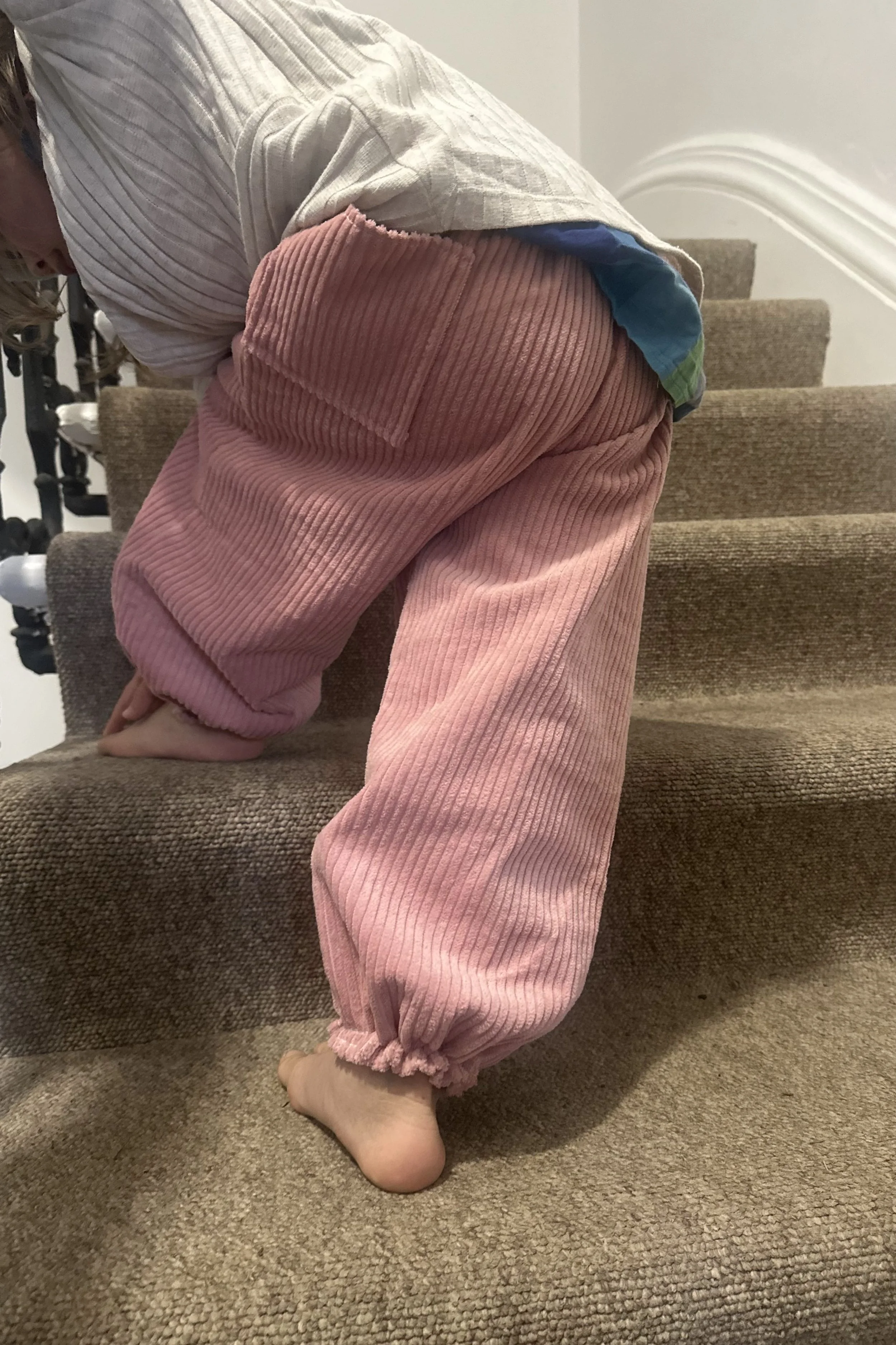 rear of child wearing organic cotton corduroy pink trousers barrel shape baggy fit oversized elastic hem, with white top, walking up staircase, highlighting pocket at back