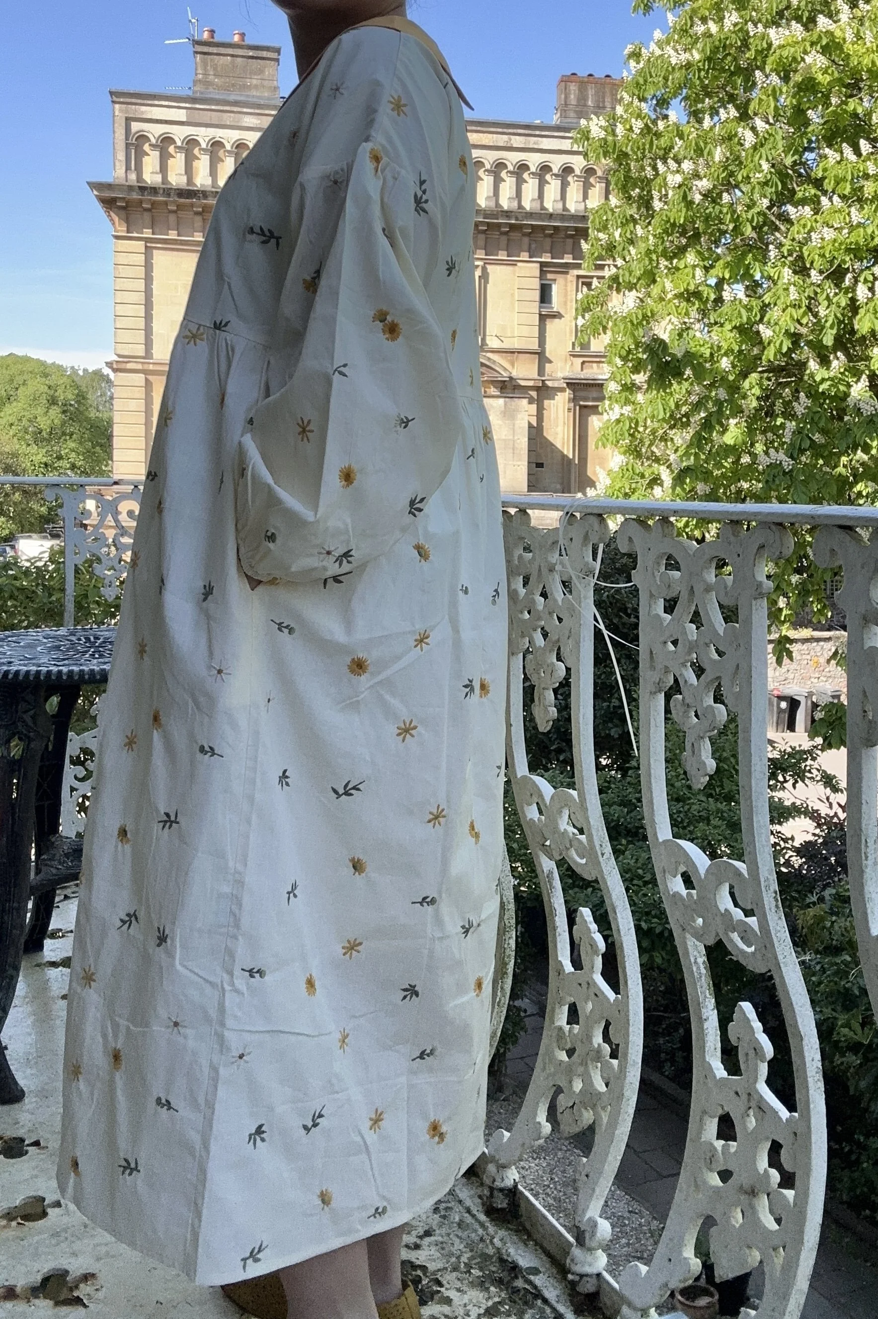 side view of woman wearing Peter Pan collar maxi summer dress dandelion embroidery standing on a balcony