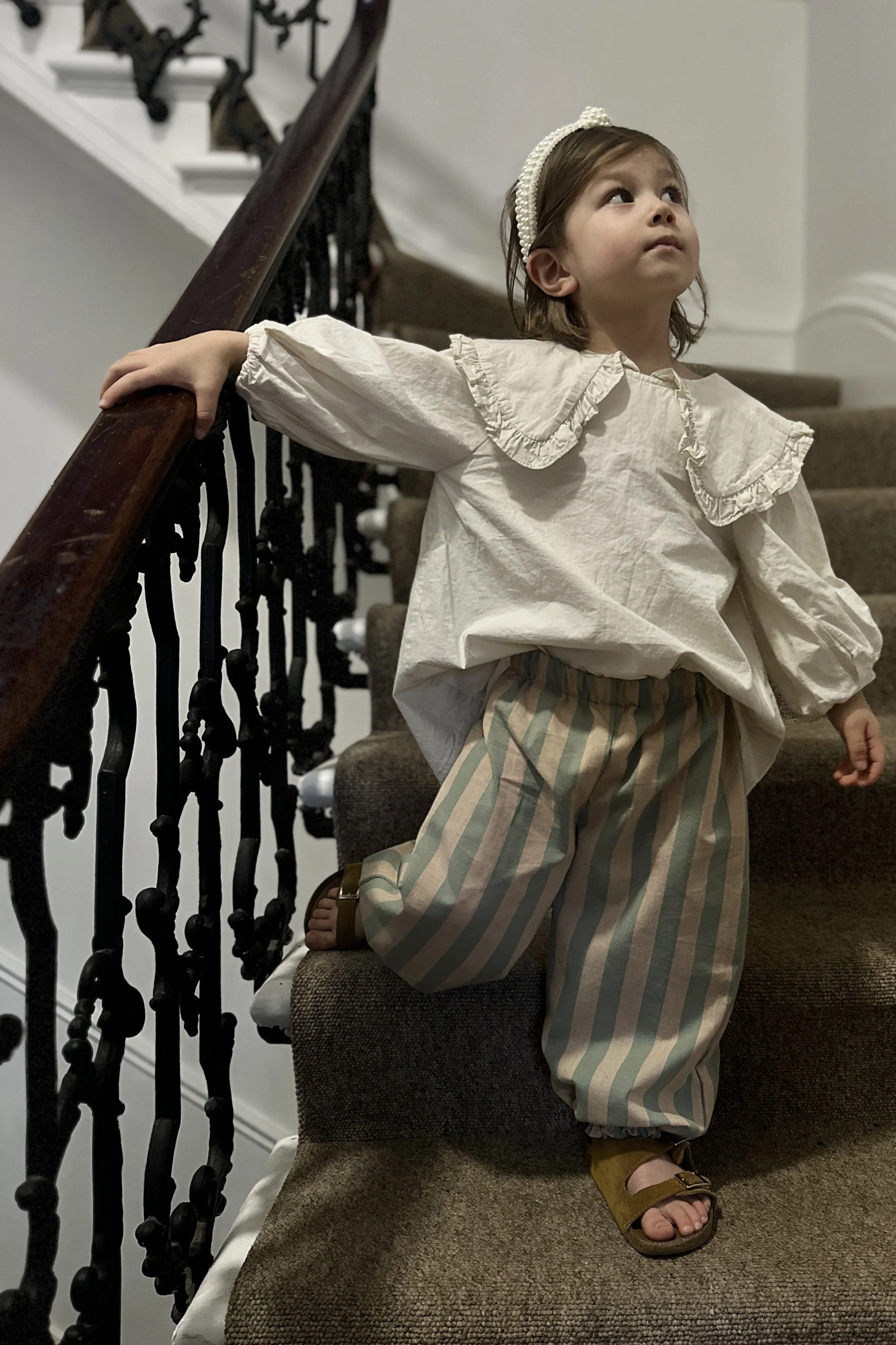 child wearing organic cotton wide stripe barrel trousers in sage green and blush pink, and a natural seeded big frill collar blouse, pearl headband, brown leather sandels