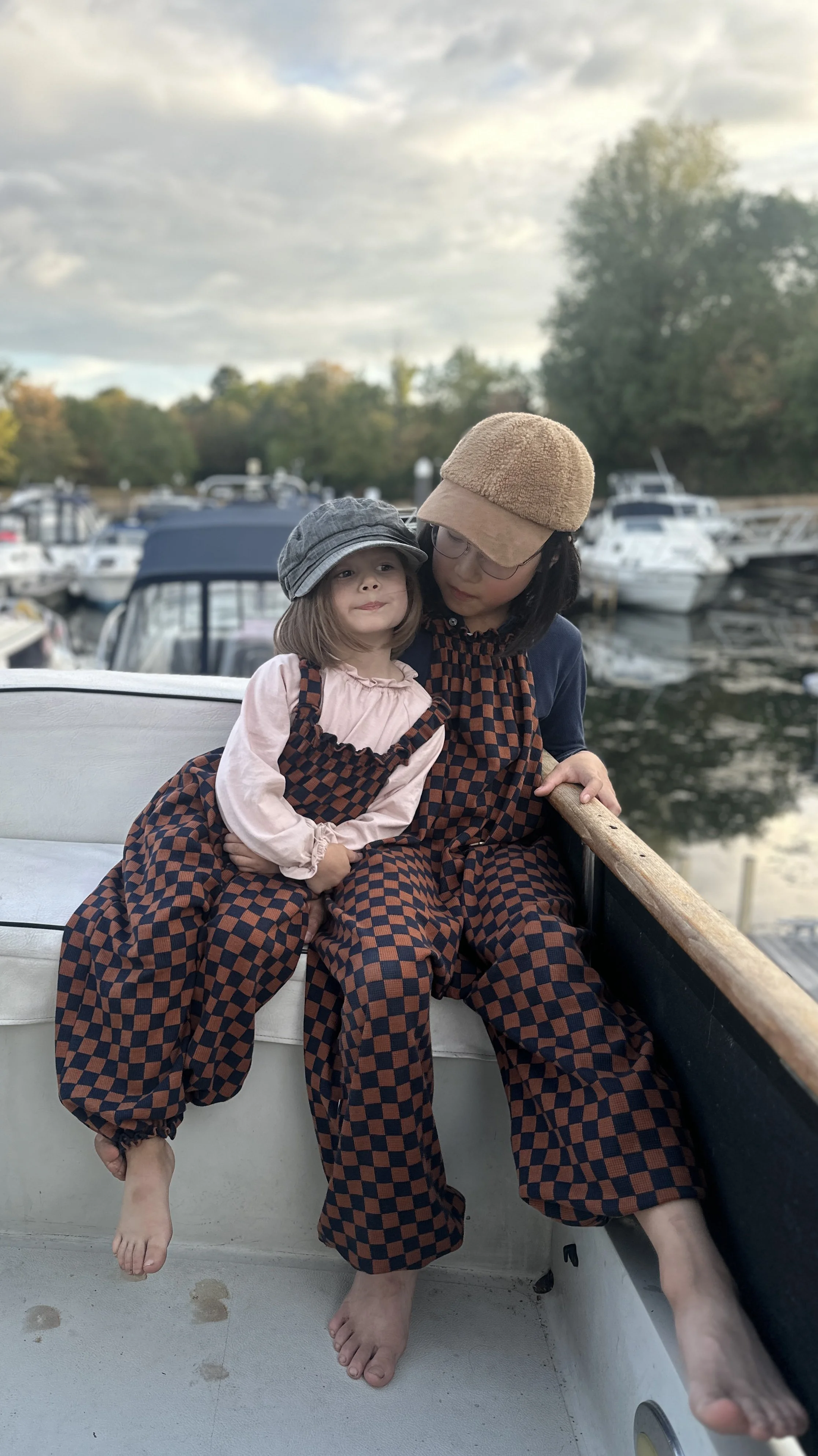 child and adult wearing pullover dungarees in checkerboard navy and rust colour, sitting on a boat at marina