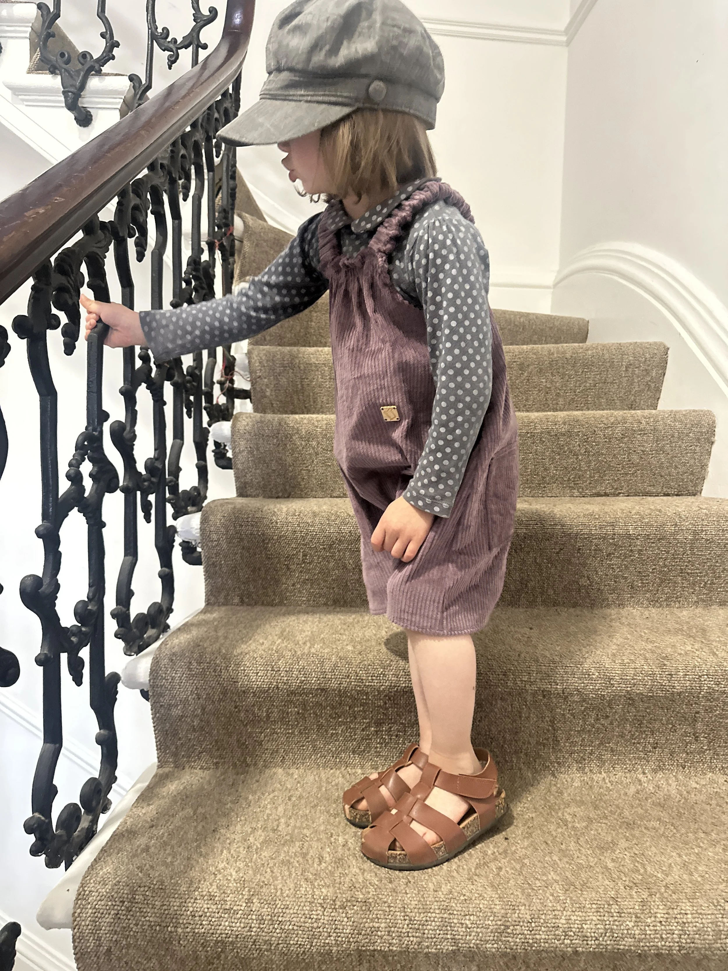 child wearing organic cotton corduroy soft purple short dungarees, layered over a grey dotty top, paired with brown sandals, grey hat