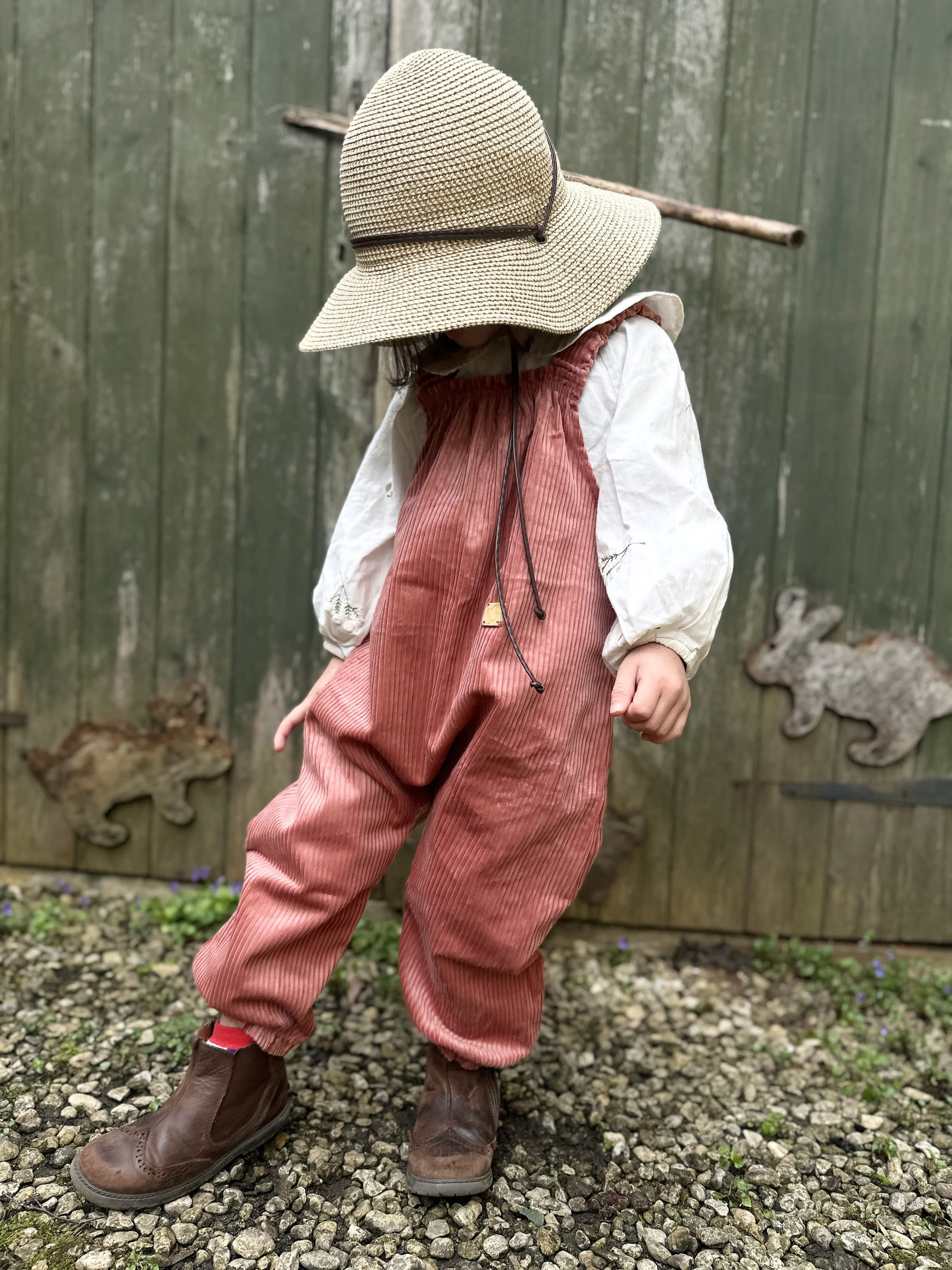 child wearing organic cotton corduroy pullover dungarees in coral colour, brown boost, sunhat, standing in front a wooden shed