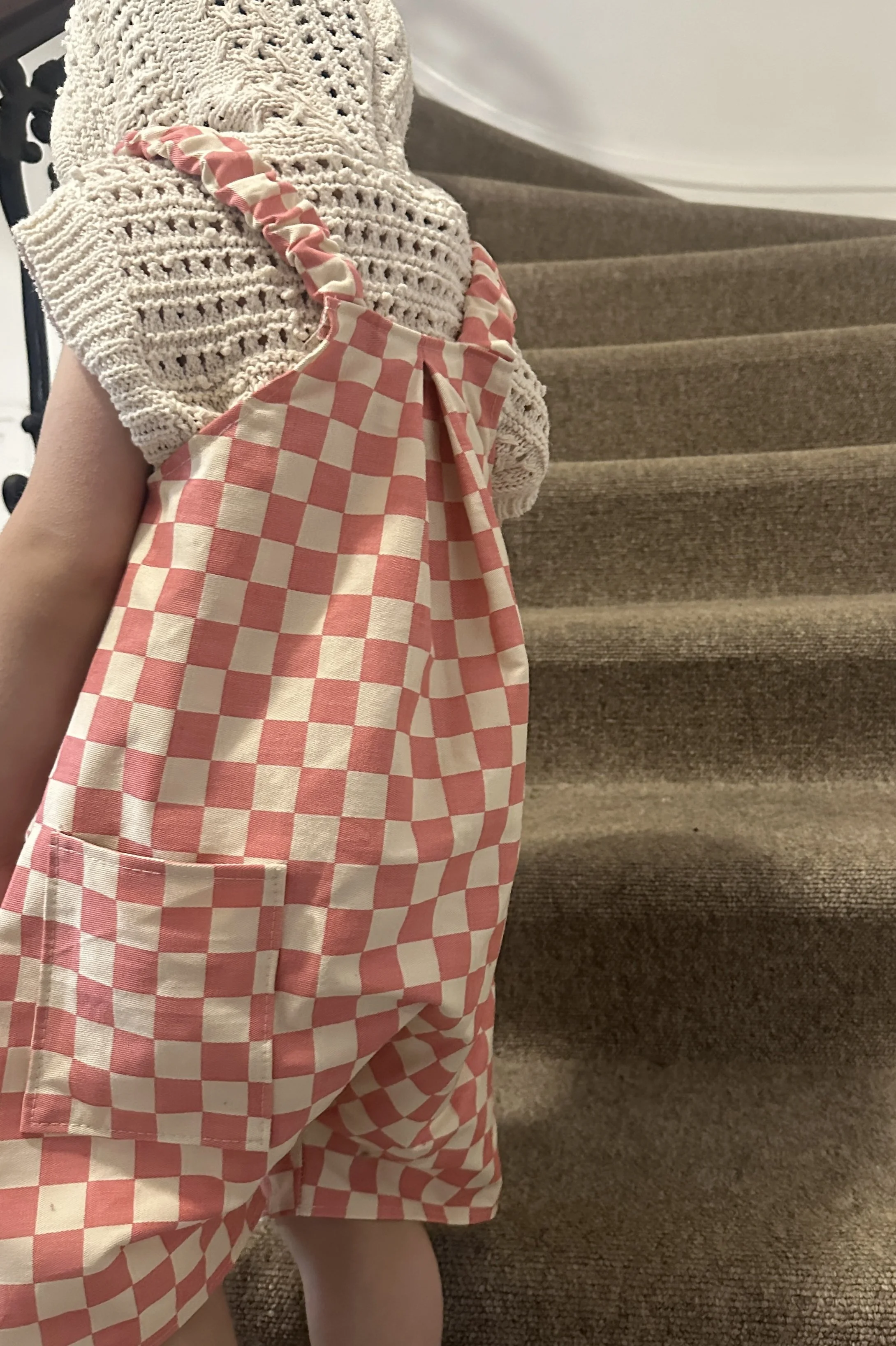 back view of child wearing organic cotton canvas pullover short dungarees in pink checkerboard colour, layered over crochet short sleeves hoodie top, brown sandals, standing on staircase