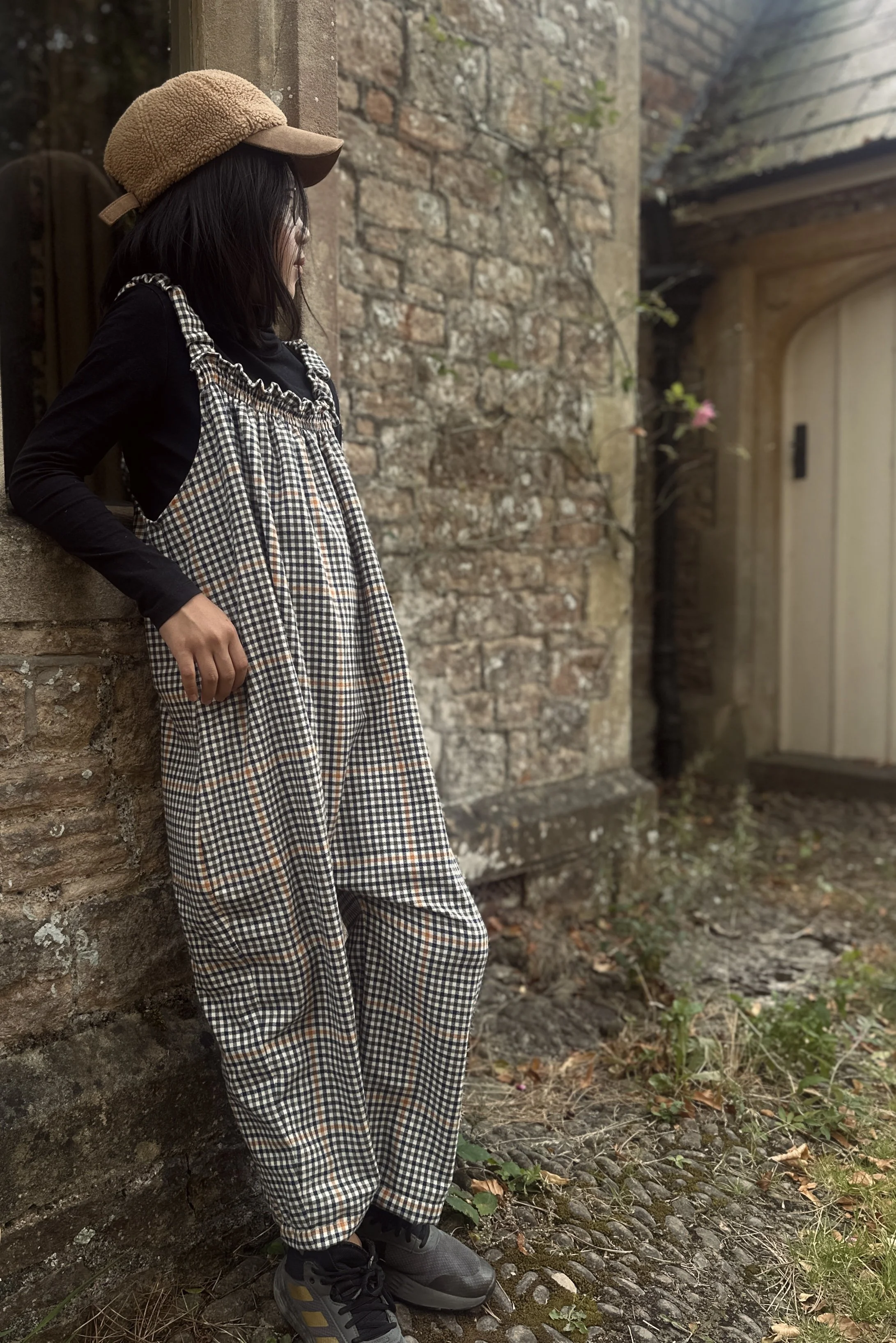 adult wearing organic cotton retro brown check dungarees layered with black top, beige cap, standing against a wall