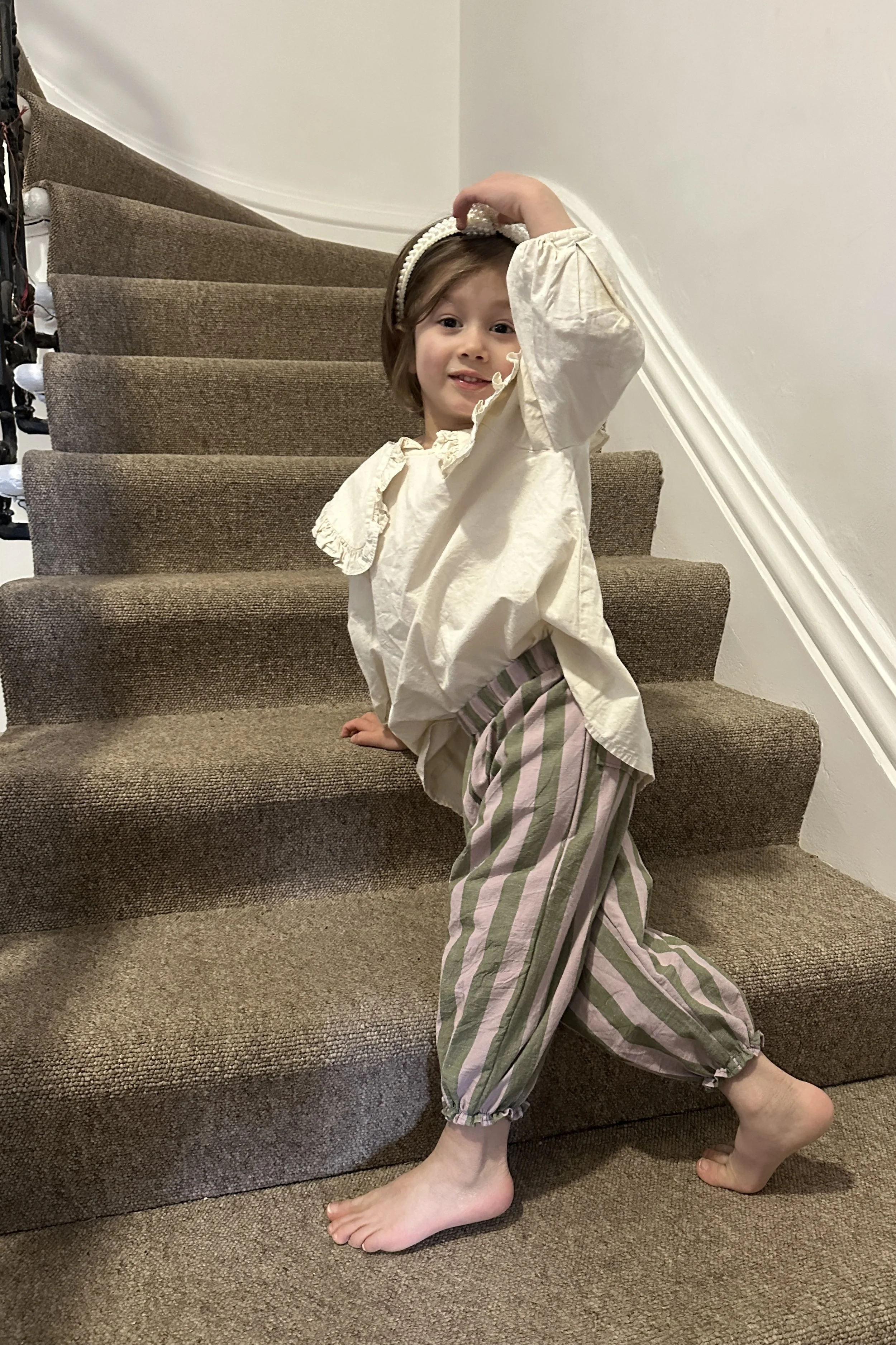 child wearing organic cotton wide stripe barrel trousers in olive green and lilac colour, paired with cream blouse with large frill collars, brown sandals, pearl hairband, standing on staircase