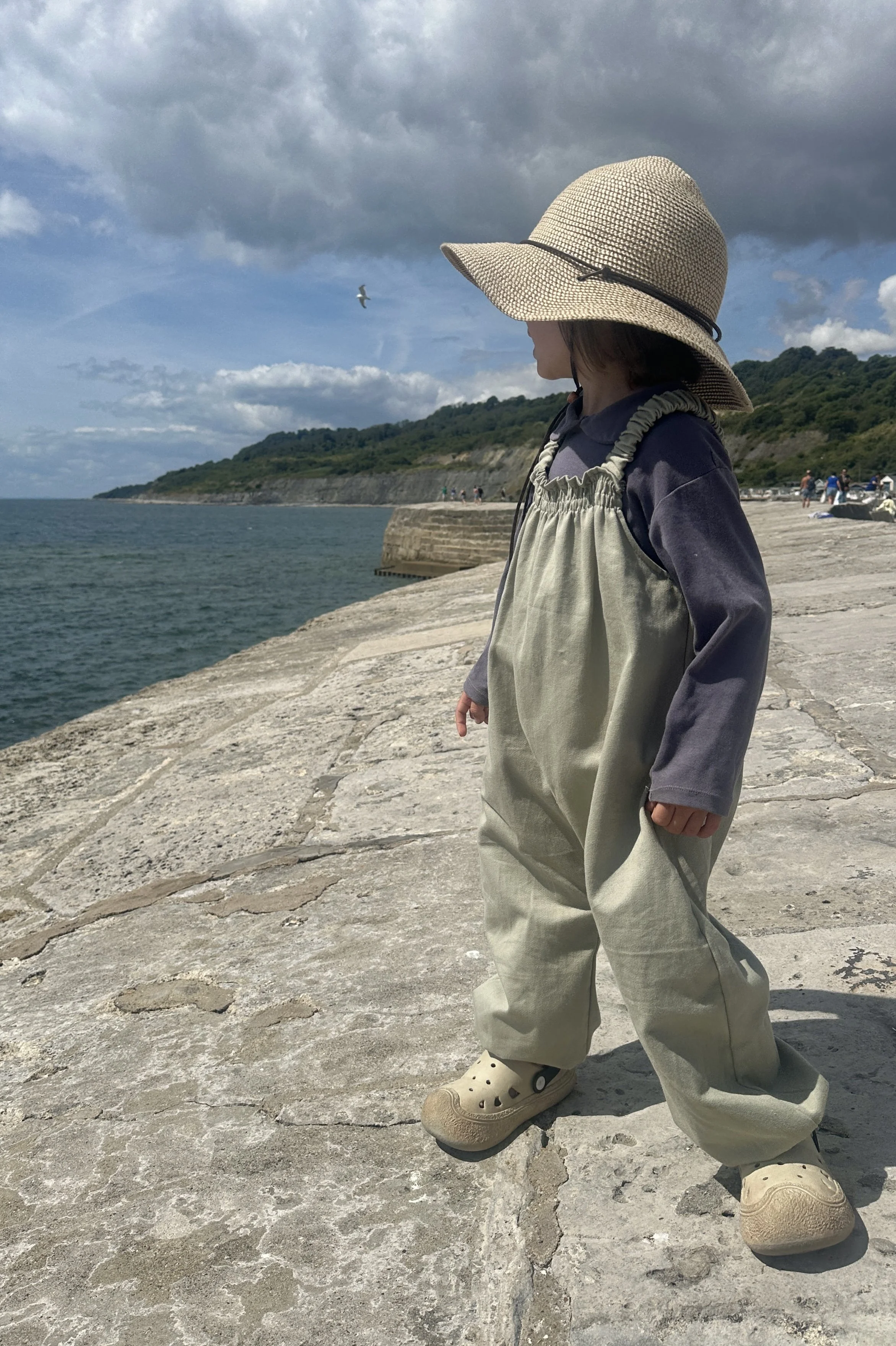 child wearing organic cotton denim light sage green pullover dungarees layered with purple top, sun hat, standing on the seaside