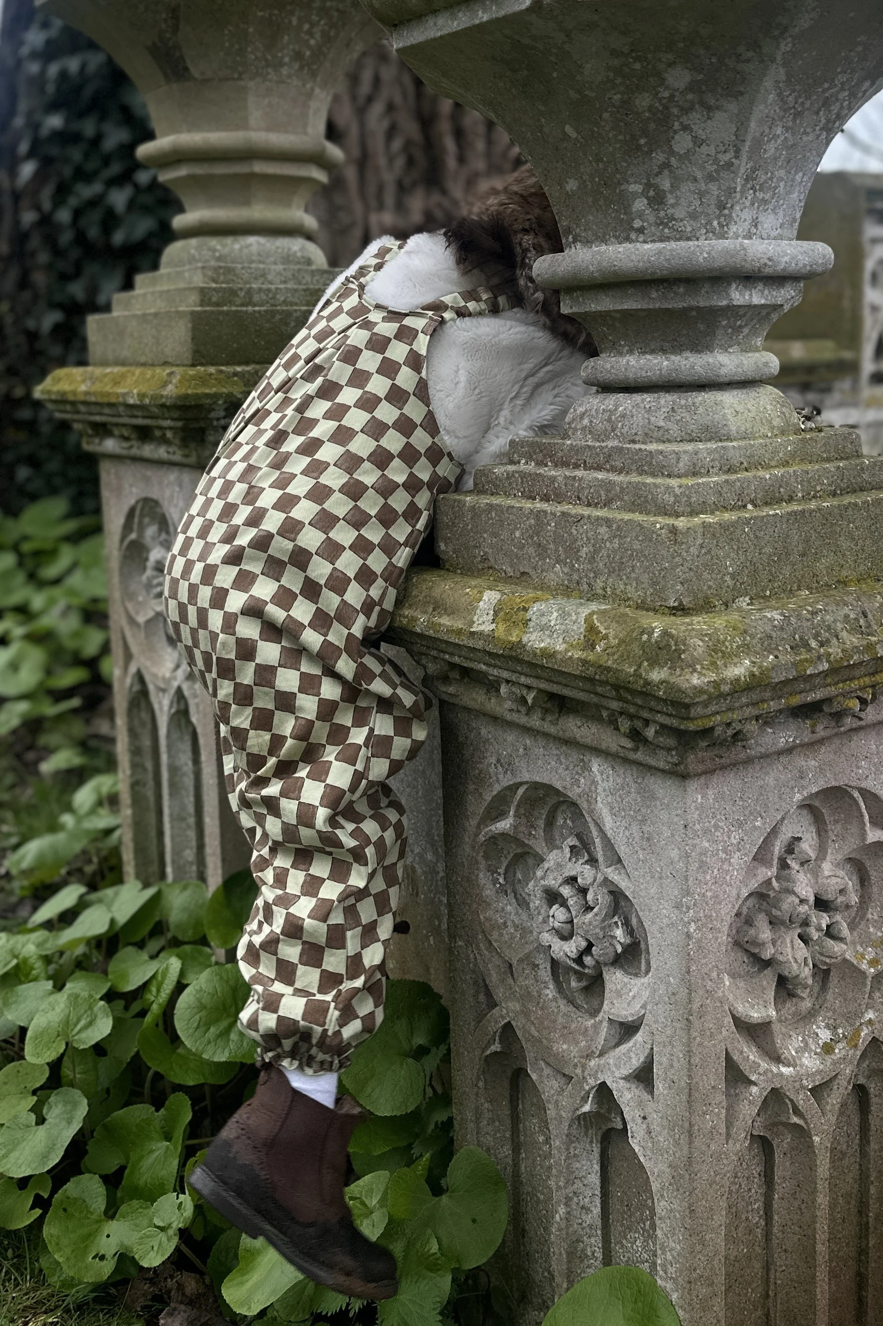 child wearing organic cotton checkerboard pullover dungarees in sage green and brown checks, brown boots, climbing a stone wall, highlighting the back