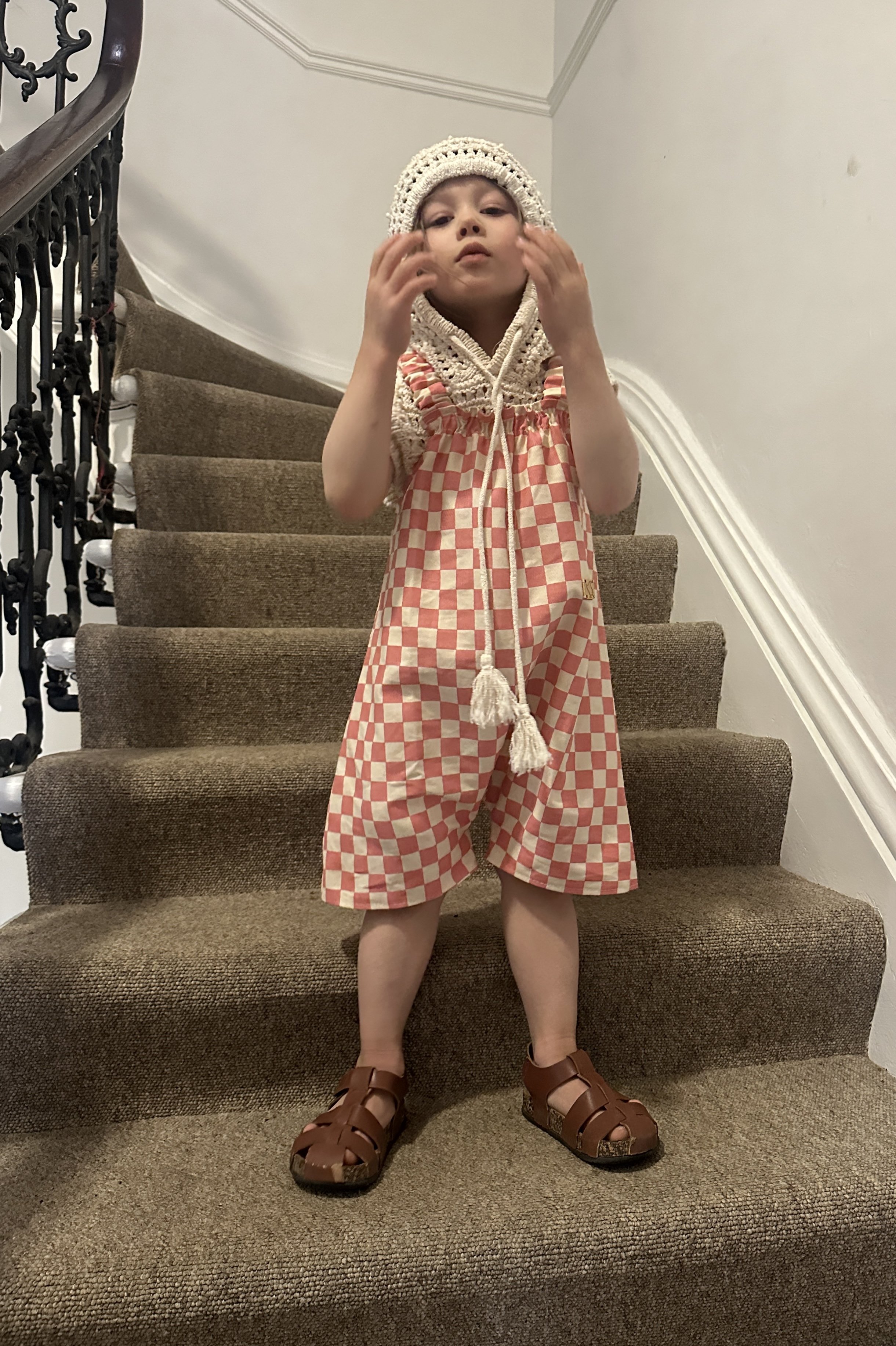 child wearing organic cotton canvas pullover short dungarees in pink checkerboard colour, layered over crochet short sleeves hoodie top, brown sandals, standing on staircase