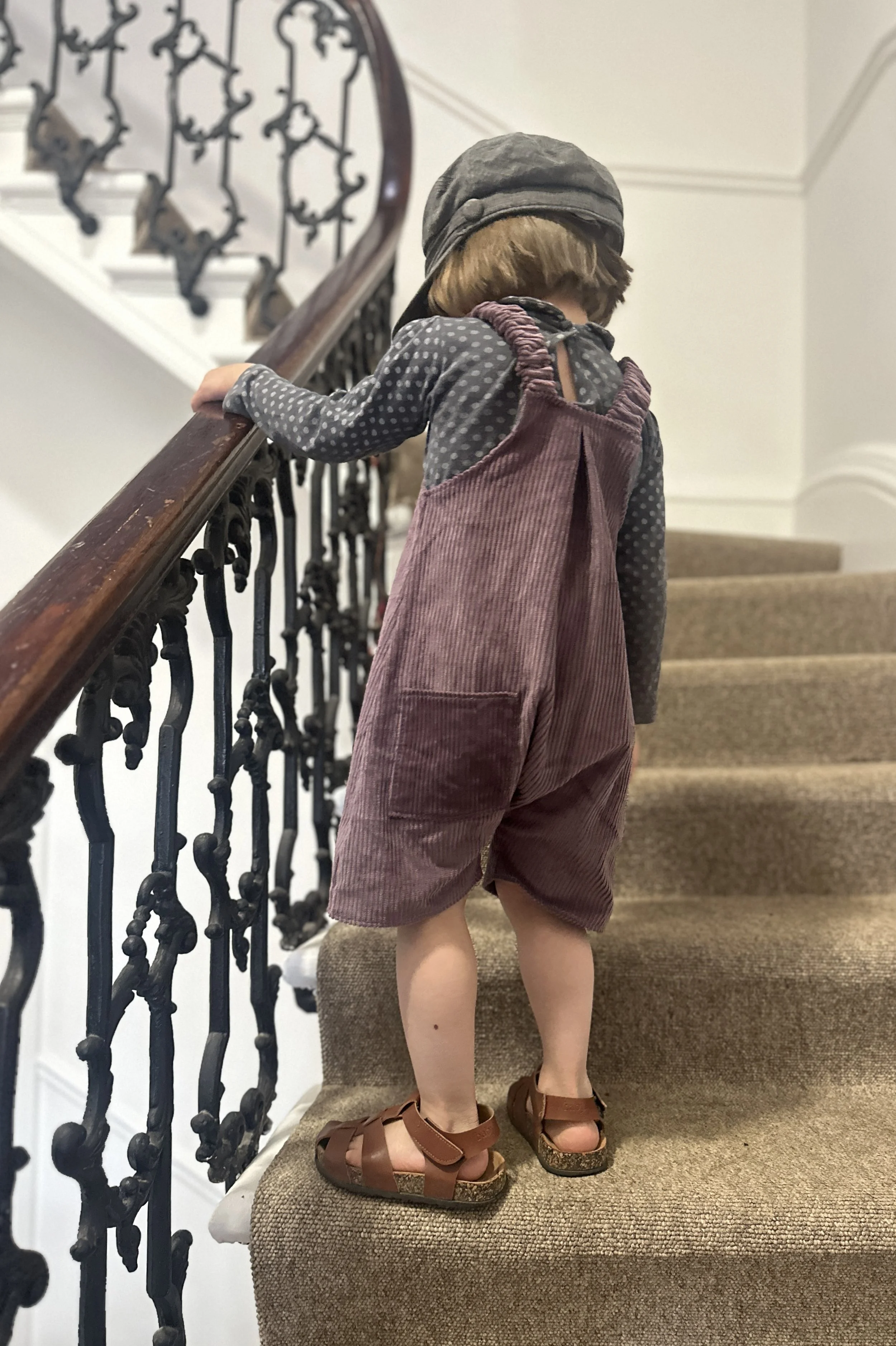 back view of child wearing organic cotton corduroy soft purple short dungarees, layered over a grey dotty top, paired with brown sandals, grey hat