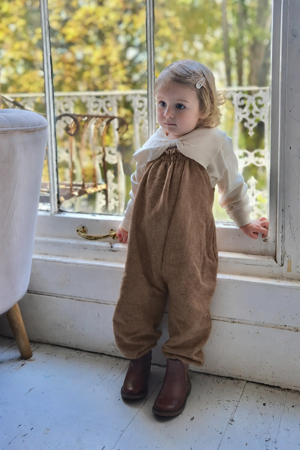 child wearing organic cotton herringbone brown dungarees layered with while collar top, standing next to a window