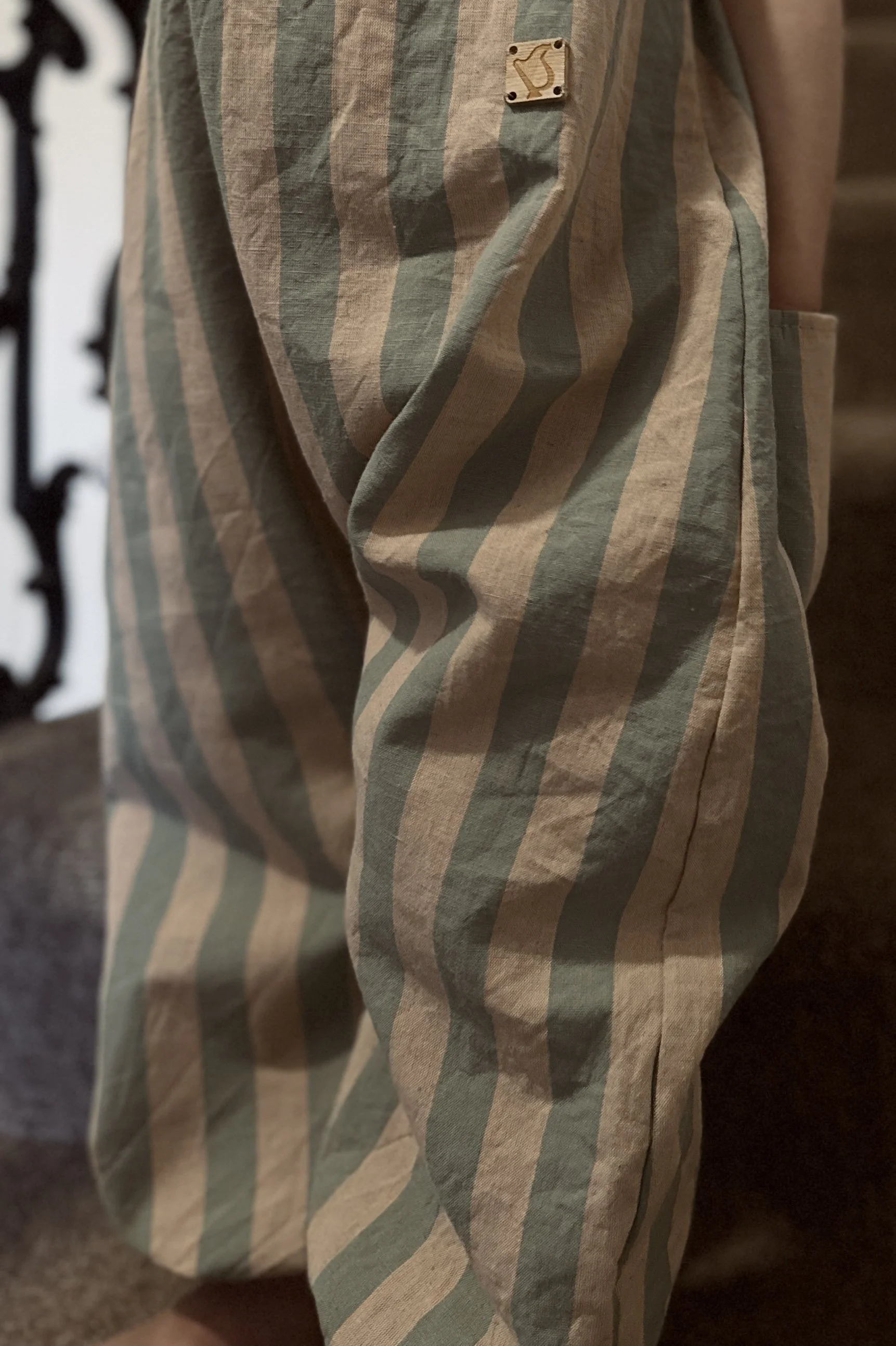 close up organic cotton wide stripe dungarees in Sage Green & Blush Pink, highlighting wooden badge and pocket at the back