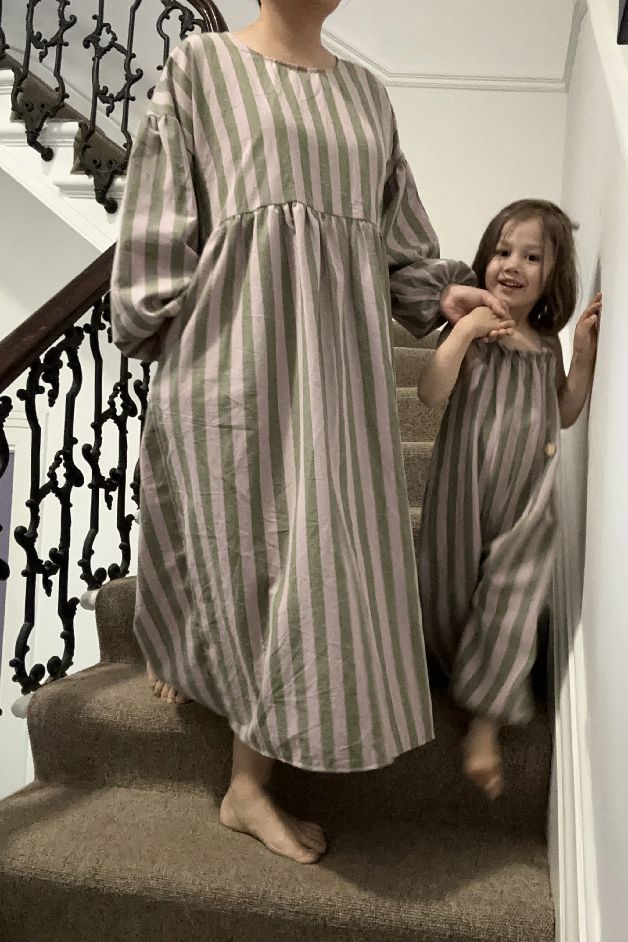 adult wearing organic cotton linen stripe olive green & lilac full length dress with balloon sleeves, with child wearing dungarees made of same fabric walking next to her on staircase