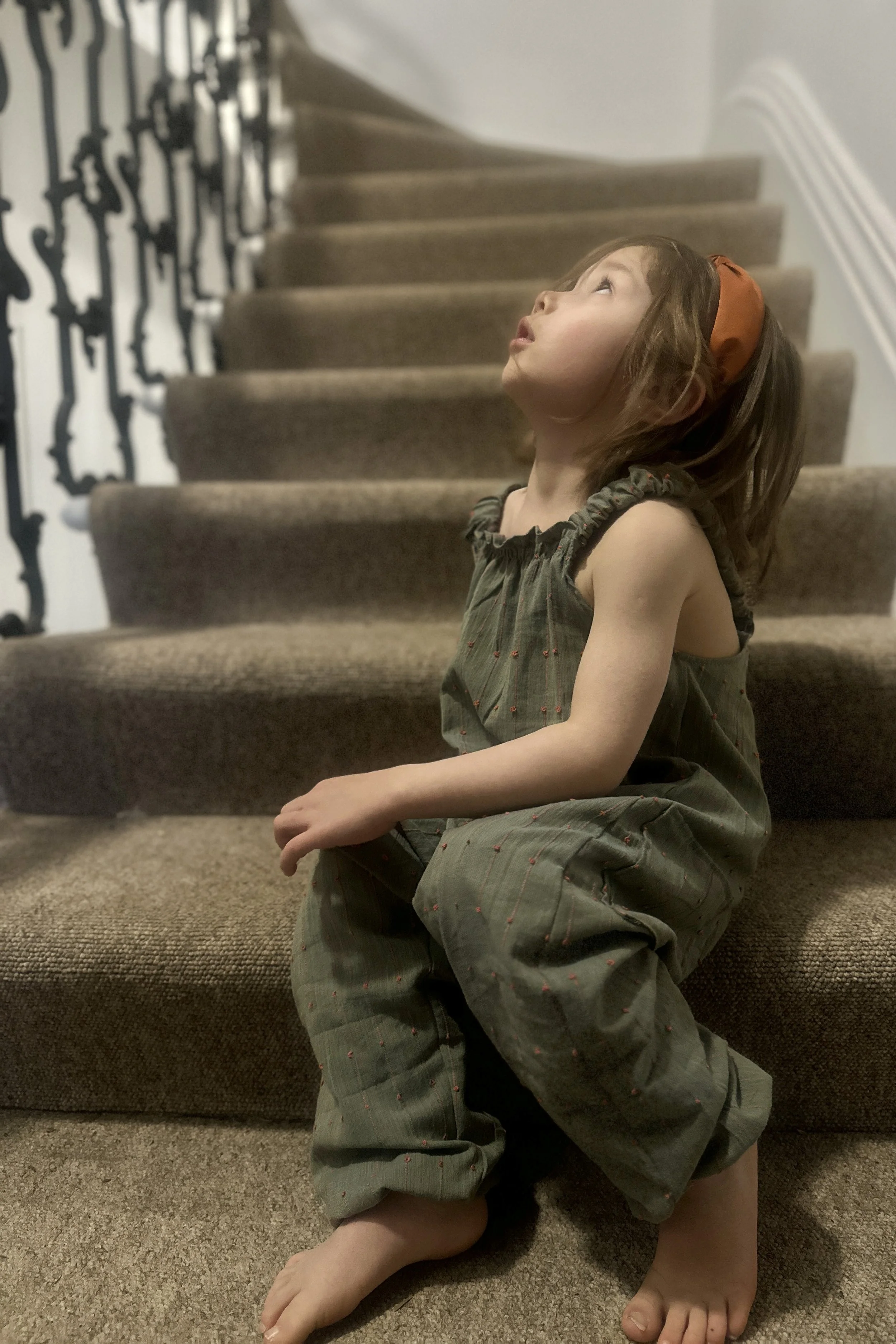 Side view of Child wearing lightweight summer pullover dungarees with coral loops on dusty sage background, with a coral colour headband, sitting on staircase