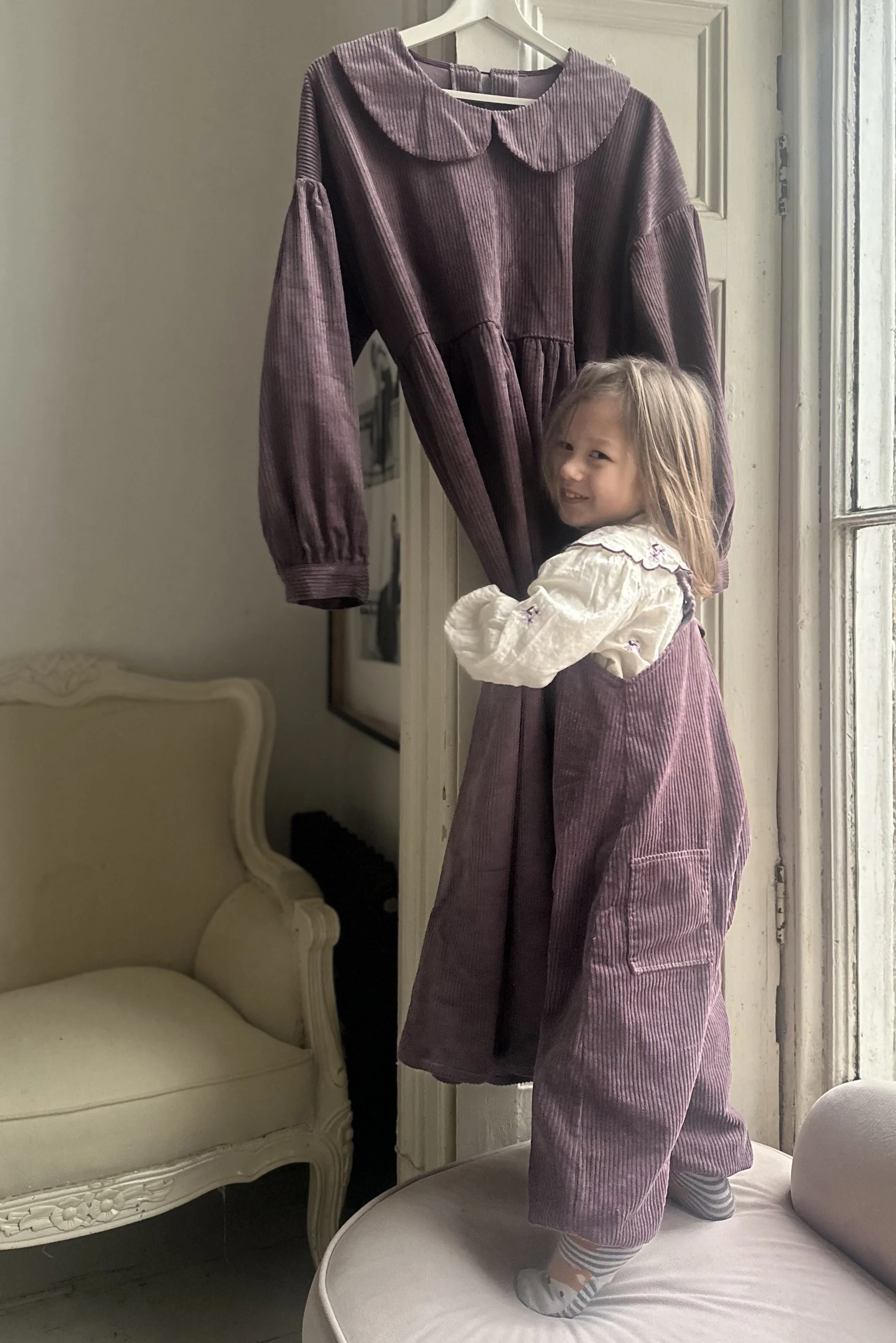 child wearing organic cotton corduroy soft purple pullover dungarees layered over a white blouse standing on a sofa holding an hanging adult long dress in same colour with Peter Pan collar