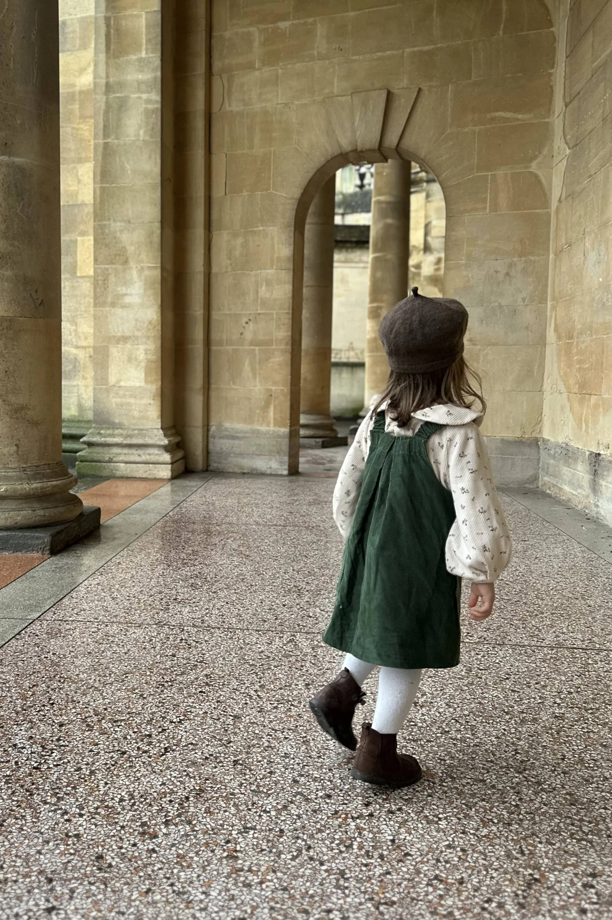rear of child wearing organic cotton needlecord dress dark green, layered over floral blouse with Peter Pan collar, a brown woolly hat and brown boots
