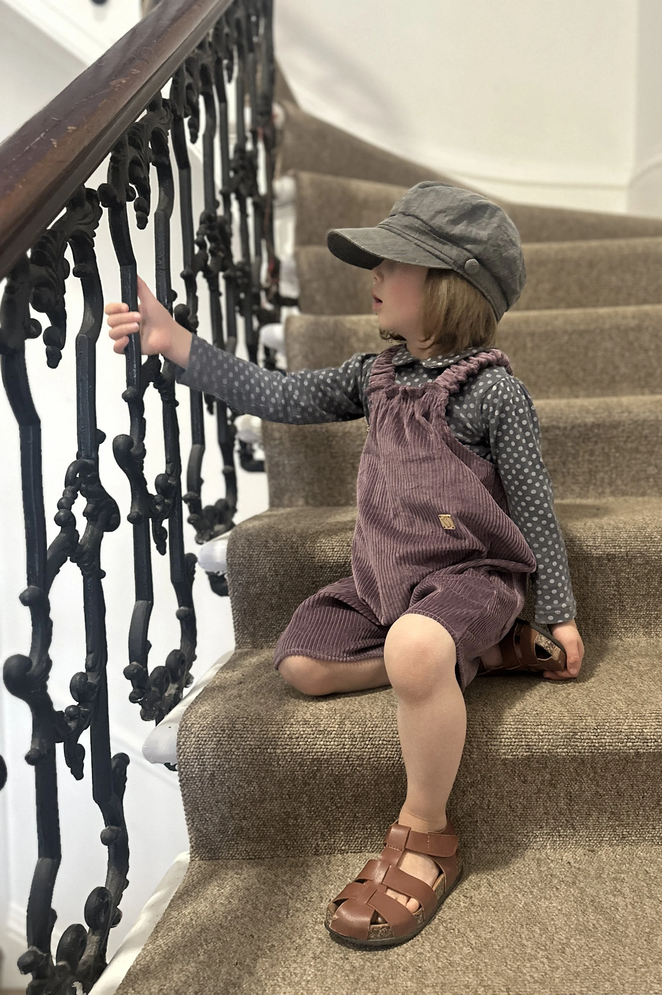 child wearing organic cotton corduroy soft purple short dungarees, layered over a grey dotty top, paired with brown sandals, grey hat, sitting on staircase